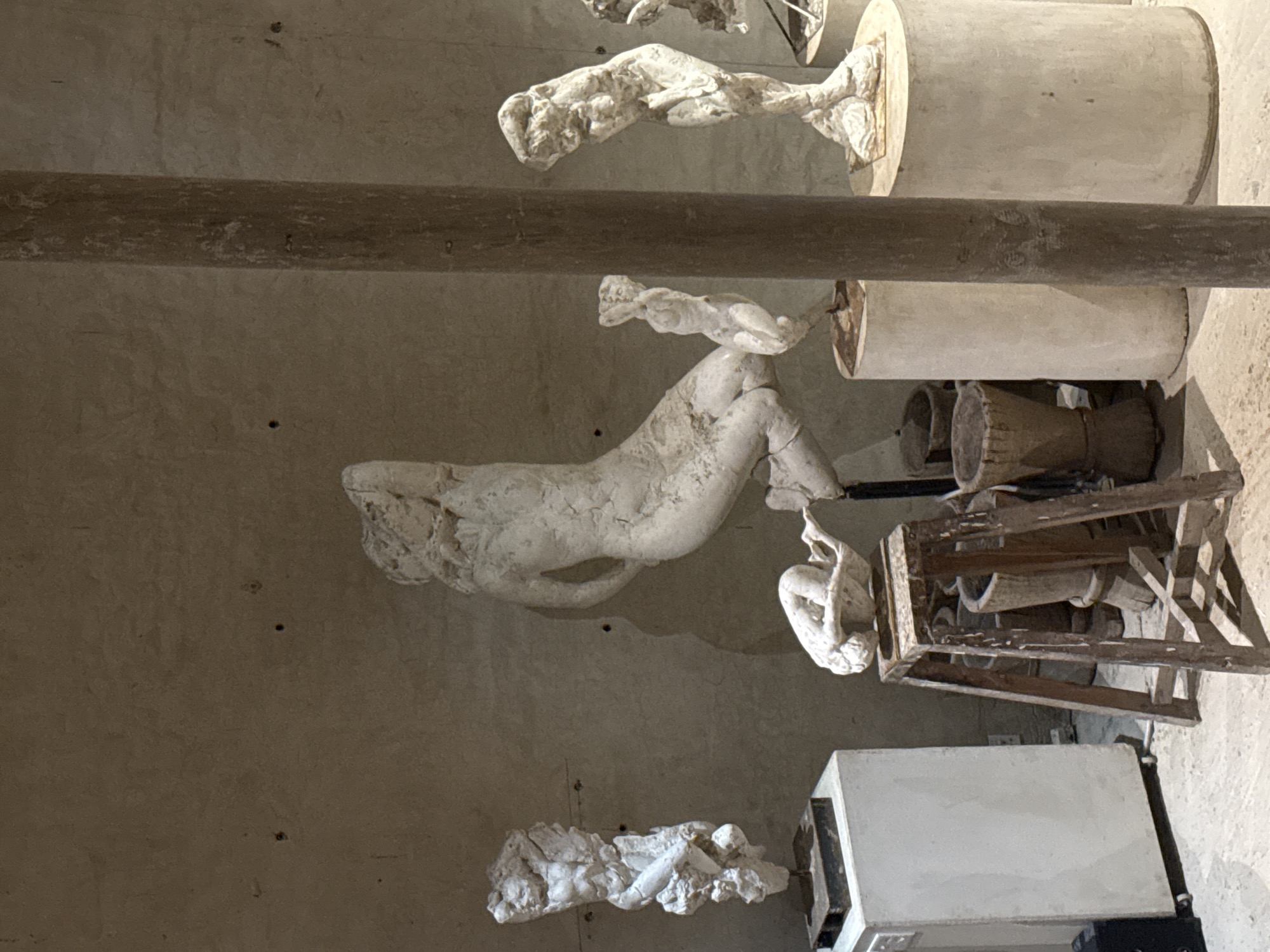 The image shows a view of a room in Stellenbosch Nu, South Africa, possibly a museum or art studio, featuring several plaster sculptures of human figures in various poses. These sculptures are mounted on cylindrical stands. A wooden beam runs horizontally across the frame, obscuring some details. The background consists of a textured wall. Other objects visible include a wooden crate and a white rectangular object, possibly a container or a display case. The lighting appears to be natural.