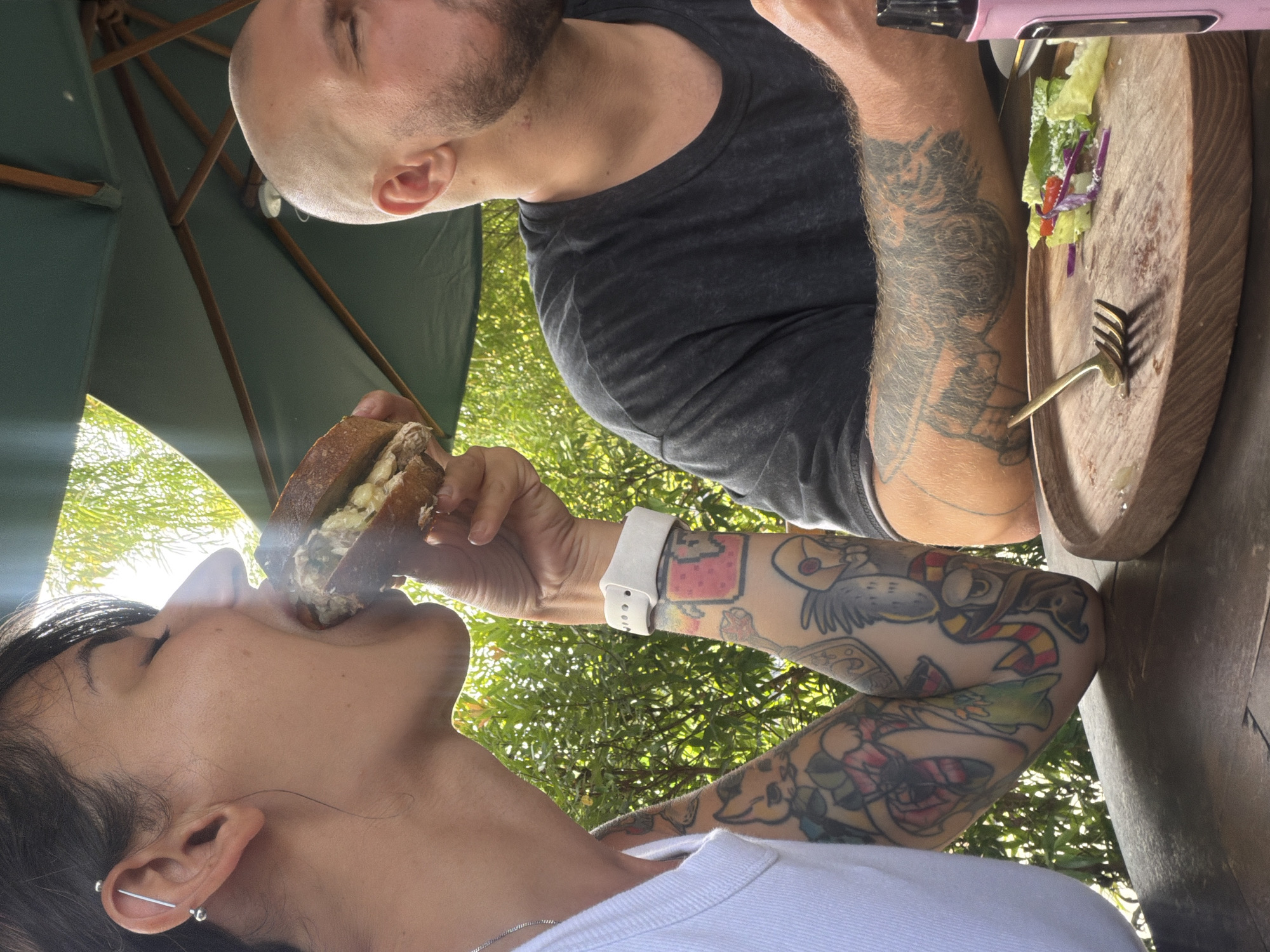 Two people are dining outdoors at a table. The person on the left, a woman with dark hair and piercings in her ear, is taking a bite of a sandwich. Her arms are covered in colorful tattoos. The person on the right, a man with a shaved head and beard, is also eating. In front of him is a wooden plate with a small salad. A pink vape pen is visible on the right side of the table. The setting appears to be a restaurant with lush green trees in the background and an umbrella overhead. The scene suggests a casual meal in a tropical environment, likely Kuta Selatan, Indonesia, given the foliage and lighting.