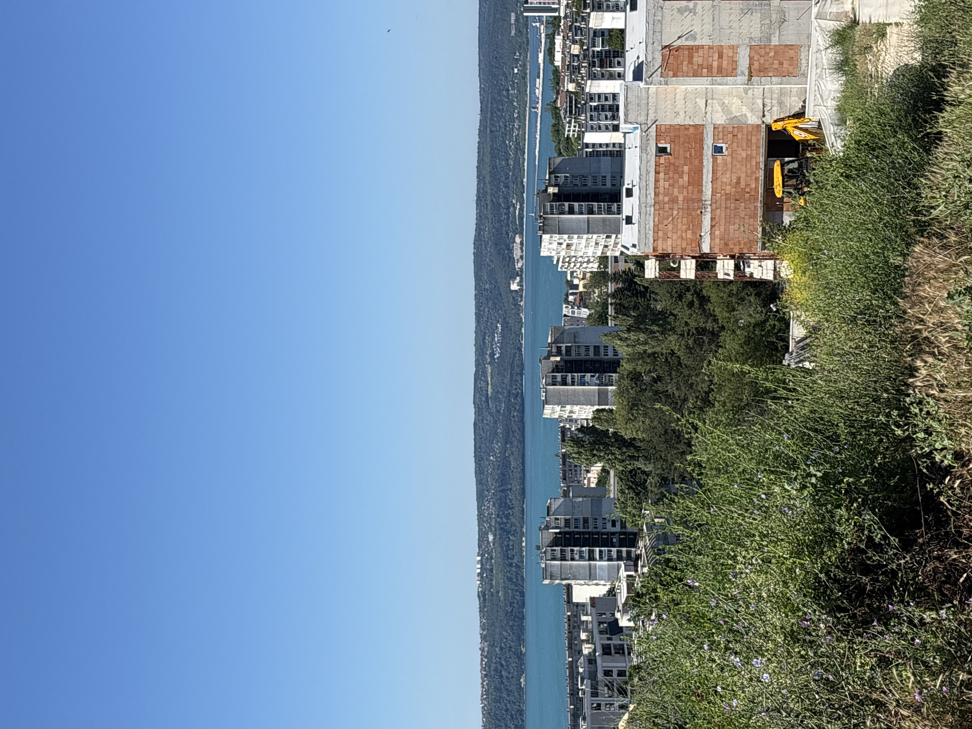 The image depicts a panoramic view of a coastal city, likely Varna, Bulgaria, under a clear blue sky. In the foreground, lush green foliage with scattered purple wildflowers and dried grasses dominates, partially obscuring a construction site. A yellow backhoe loader is visible on the construction site, which features buildings under various stages of completion, including a structure with exposed red brickwork and concrete framing.
Beyond the immediate foreground, a dense cluster of apartment buildings, primarily gray and white, rises towards the horizon. These buildings exhibit a consistent architectural style, with repetitive window patterns and balconies. Further in the distance, a wide expanse of turquoise water, likely the Black Sea, stretches across the frame, bordered by a forested coastline and a low-lying hill with scattered structures. The distant shoreline is dotted with buildings and hints at the city's urban sprawl extending along the coast. The overall scene suggests a sunny day in a developing urban area adjacent to a significant body of water.