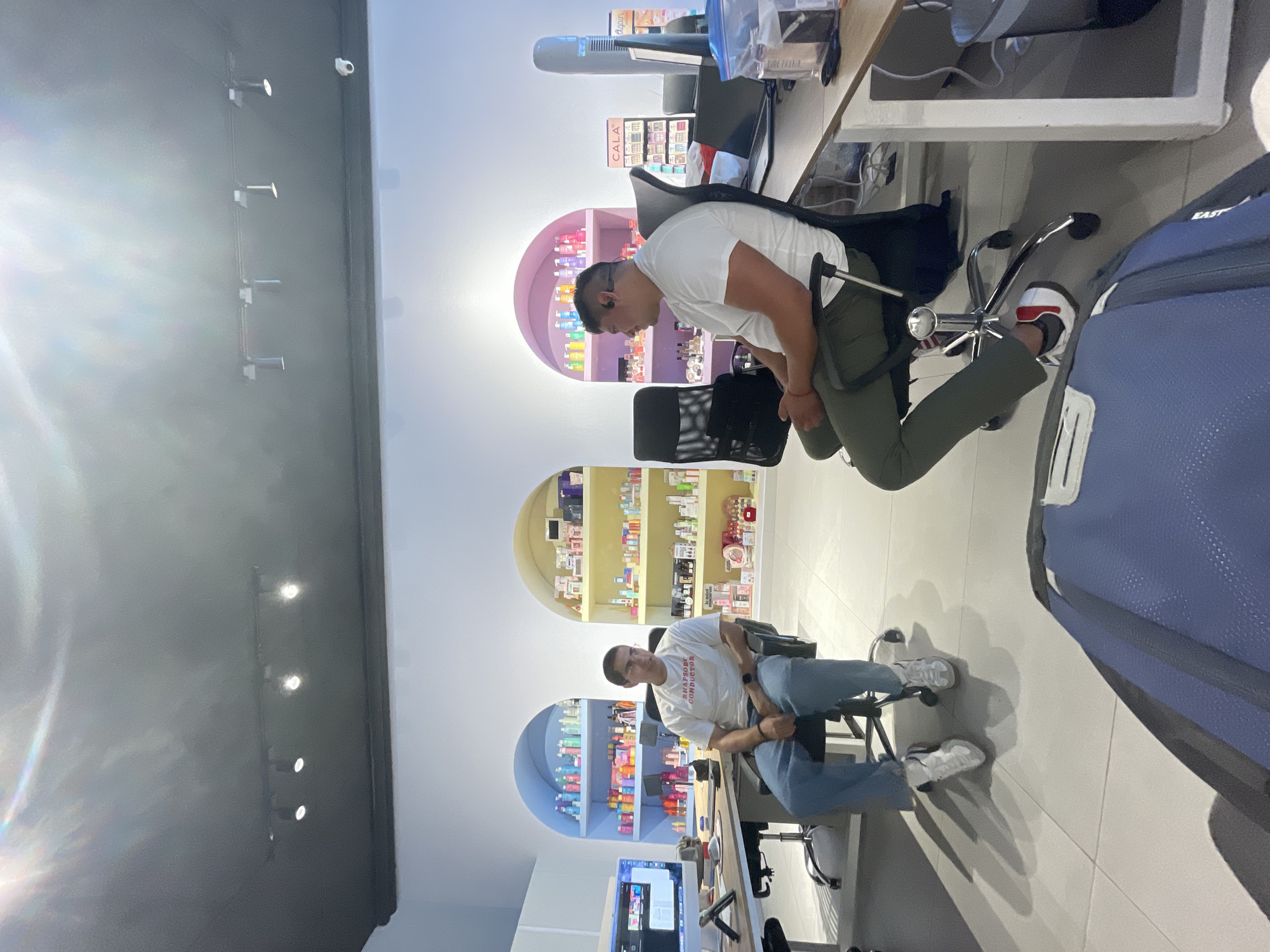A commercial space is captured in this medium, indoor shot, rotated 90 degrees clockwise. Two men are seated at desks, in front of arched shelves lined with colorful retail products, which may indicate a retail store.
The first man, at the top of the image, wears a white t-shirt and olive-green pants. He is seated in an office chair, angled away from the camera. The second man, at the bottom of the image, is dressed in a white t-shirt and blue jeans. He is also seated in an office chair.
A blue backpack is situated in the lower right corner. Computer monitors and office equipment are scattered on the desks. Spotlights are visible on the ceiling.
Based on the retail environment with arched shelves and cosmetic products, it is likely that this location is in Mexico City, Mexico.