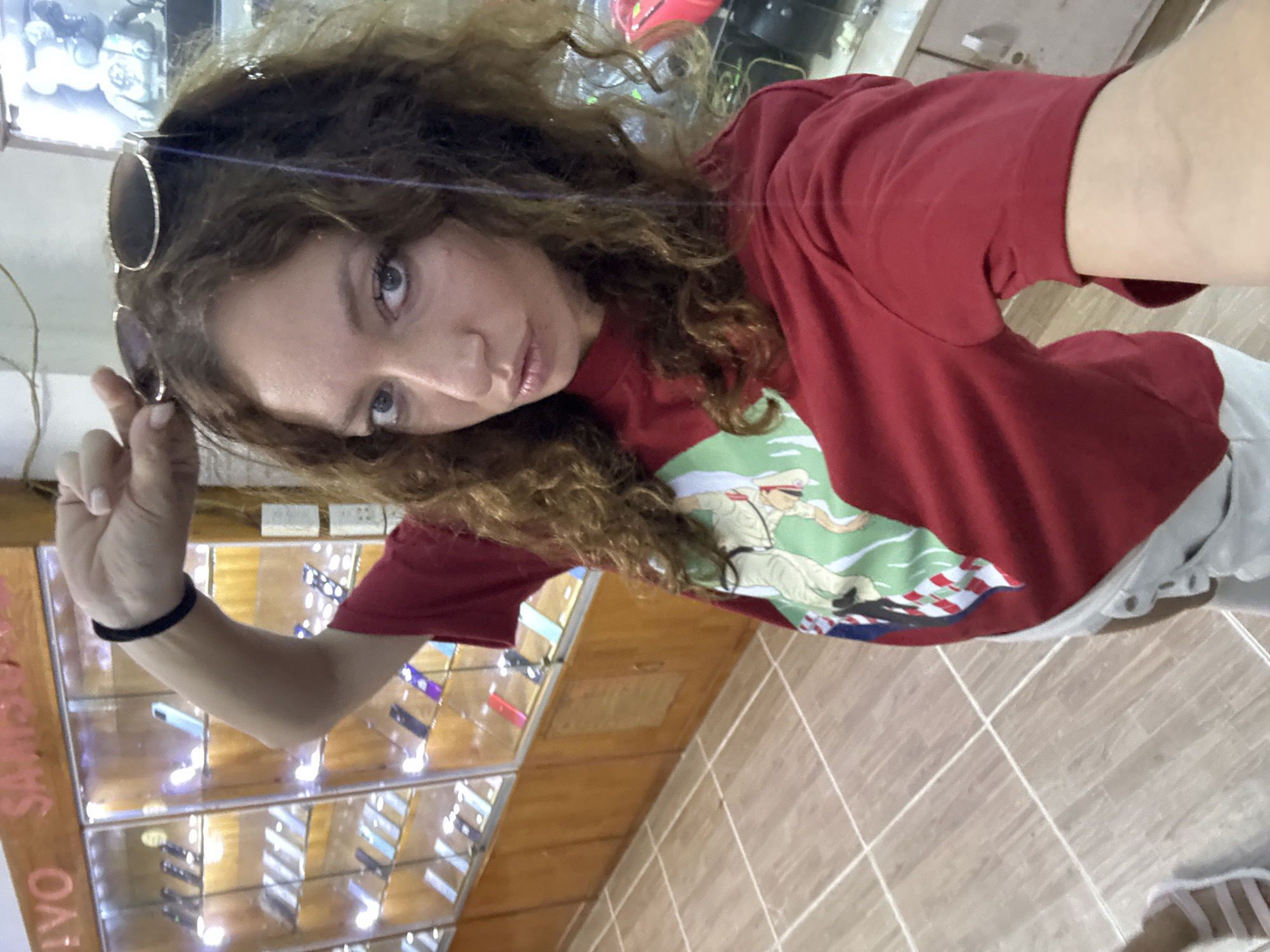 A young woman with curly hair and sunglasses in her hand takes a selfie inside a shop. She wears a red T-shirt with a graphic print. Behind her is a glass display case filled with small objects. The shop is located in the Du Già Commune, Vietnam. The floor is tiled.