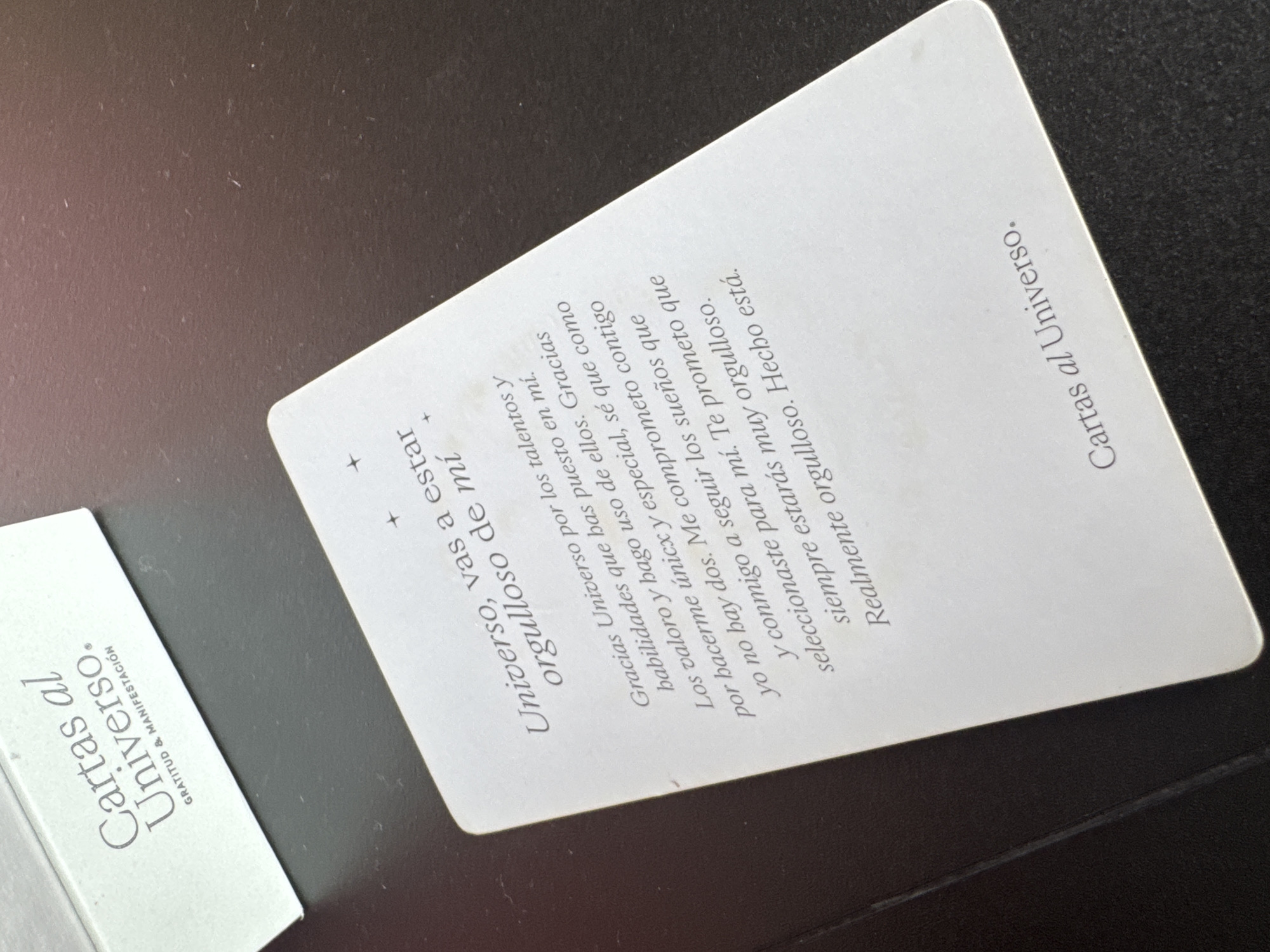 An overhead shot captures two "Cartas al Universo" (Letters to the Universe) cards and/or packaging elements resting on a dark, flat surface, possibly a table or desk, in San Bernardino Tlaxcalancingo, Mexico.
The main subject is a white, rectangular card positioned diagonally, occupying the right and central portions of the frame. The card displays text in Spanish, appearing to be a personal affirmation or manifestation statement. The title "Cartas al Universo." is visible at the top right of this card. The main body of text reads: "Universo, vas a estar orgulloso de mí +" (Universe, you will be proud of me +). Below this, a paragraph continues: "Gracias Universo por los talentos y habilidades que has puesto en mí. Los valoro y hago uso de ellos. Gracias por hacerme únicx y especial, sé que como yo no hay dos. Me comprometo contigo y conmigo a seguir los sueños que seleccionaste para mí. Te prometo que siempre estarás muy orgulloso. Realmente orgulloso. Hecho está." (Thank you Universe for the talents and abilities you have given me. I value them and make use of them. Thank you for making me unique and special, I know there's no one like me. I commit to you and to myself to follow the dreams you selected for me. I promise you will always be very proud. Truly proud. It is done.) The use of "únicx" indicates gender-inclusive language.
To the left, a partial view of another white card or the packaging box for the cards is visible, also tilted. On this item, the prominent text "Cartas al Universo." is seen, along with "GRATITUD & MANIFESTACIÓN" (Gratitude & Manifestation) below it.
The dark background surface shows minor dust particles, indicating a casual, everyday setting. The lighting suggests an indoor environment, likely daytime, without harsh shadows. The items appear to be tools for spiritual or self-help practices.