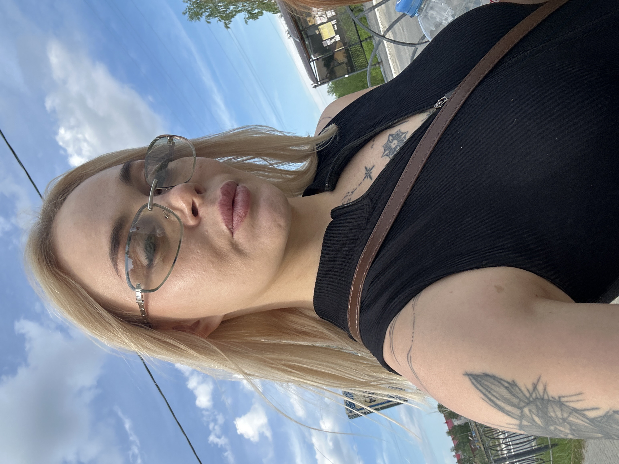 A fair-skinned female with long, straight blonde hair, parted on her left side and appearing slightly windblown, takes a selfie outdoors under a clear blue sky with scattered white clouds. She wears large, light-tinted, rimless sunglasses. Her lips are full and have a light pink cosmetic application. She is dressed in a black, ribbed, high-collar, sleeveless top with a partial front zipper. A brown strap, consistent with a shoulder bag, crosses diagonally over her chest. Tattoos are visible: a linear, geometric design on her left upper arm; a thin, dotted line over her left shoulder; and two distinct designs on her chest, a stylized star or cross above a concentric mandala-like pattern, visible through the unzipped collar. In the background, a utility wire traverses the mid-left frame, above indistinct buildings and a dark metal fence. To the right, green foliage, a dark-roofed shelter, and the partial view of a second individual with reddish-auburn hair are present. A clear plastic water bottle with a blue cap is visible in the lower right foreground. The scene is brightly illuminated by natural daylight.
