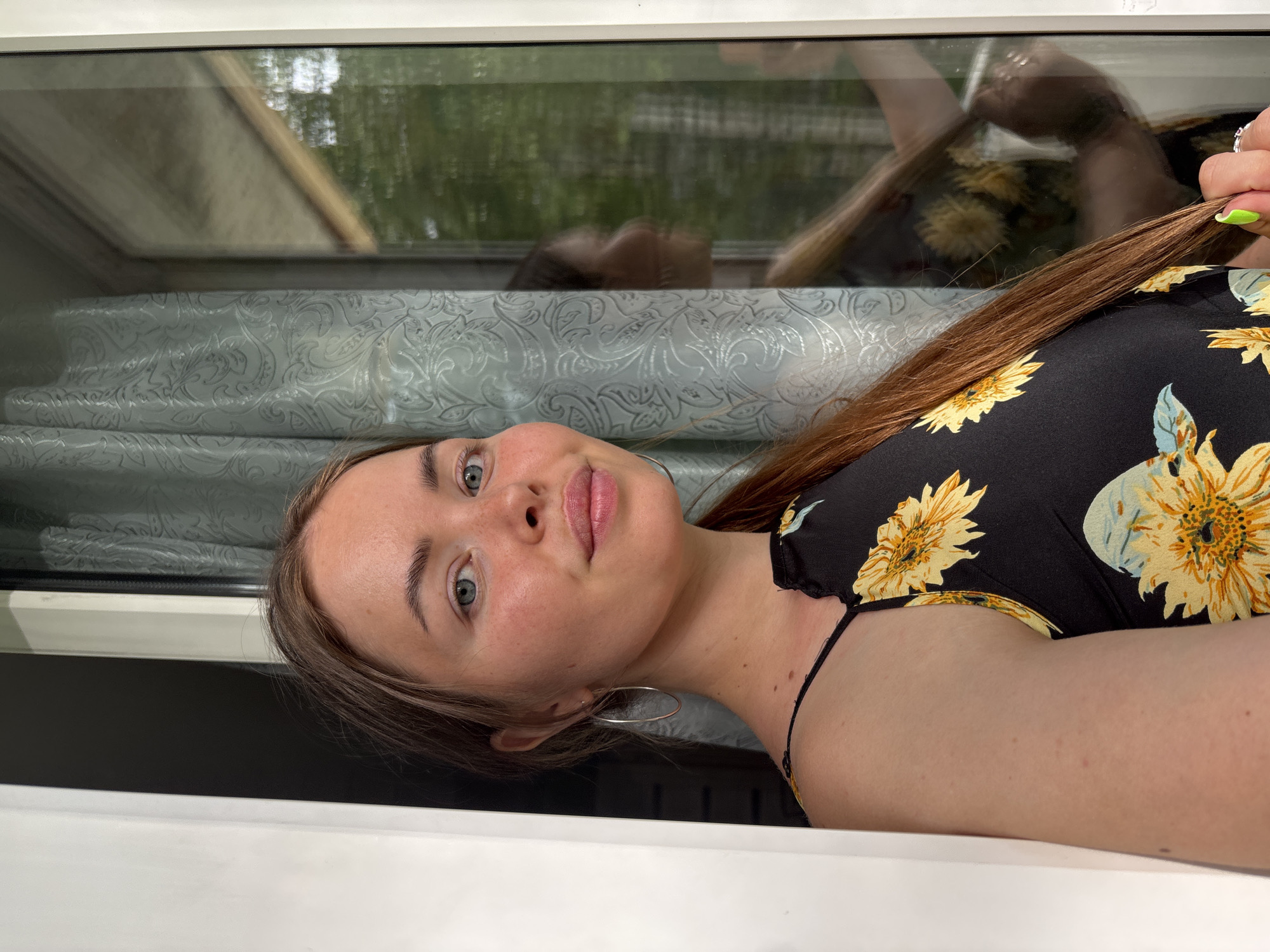 A young woman is lying down, likely on a white surface. She is wearing a black top with sunflower prints. Her hair is long and brown. She is wearing a silver hoop earring in her left ear. Behind her is a window with a reflection of herself and trees. The reflection suggests she may be in an interior space, potentially near a park or wooded area. There is no specific information in the image to determine the location in the city of Izhevsk, Russia.