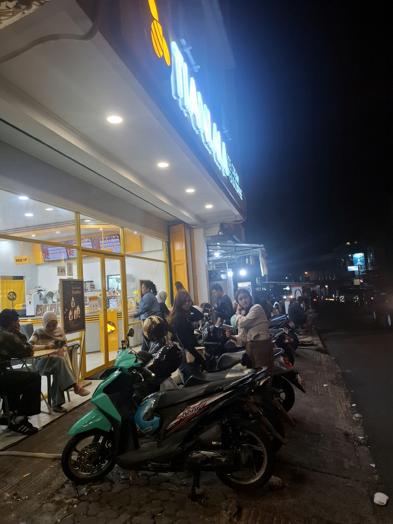 This is a nighttime photograph of a brightly lit outdoor seating area and storefront, presumably located in Cianjur, Indonesia. A prominent neon sign above the establishment reads "MAJU JAYA." Several people are seated at tables outside the storefront, which features a large glass window displaying menu boards and interior lighting. A "PICK UP" sign is visible near the entrance. A variety of motorcycles are parked along the sidewalk in front of the seating area. The street to the right of the sidewalk is dark, with some distant building lights and vehicle headlights visible. The overall scene depicts a casual, commercial setting at night.