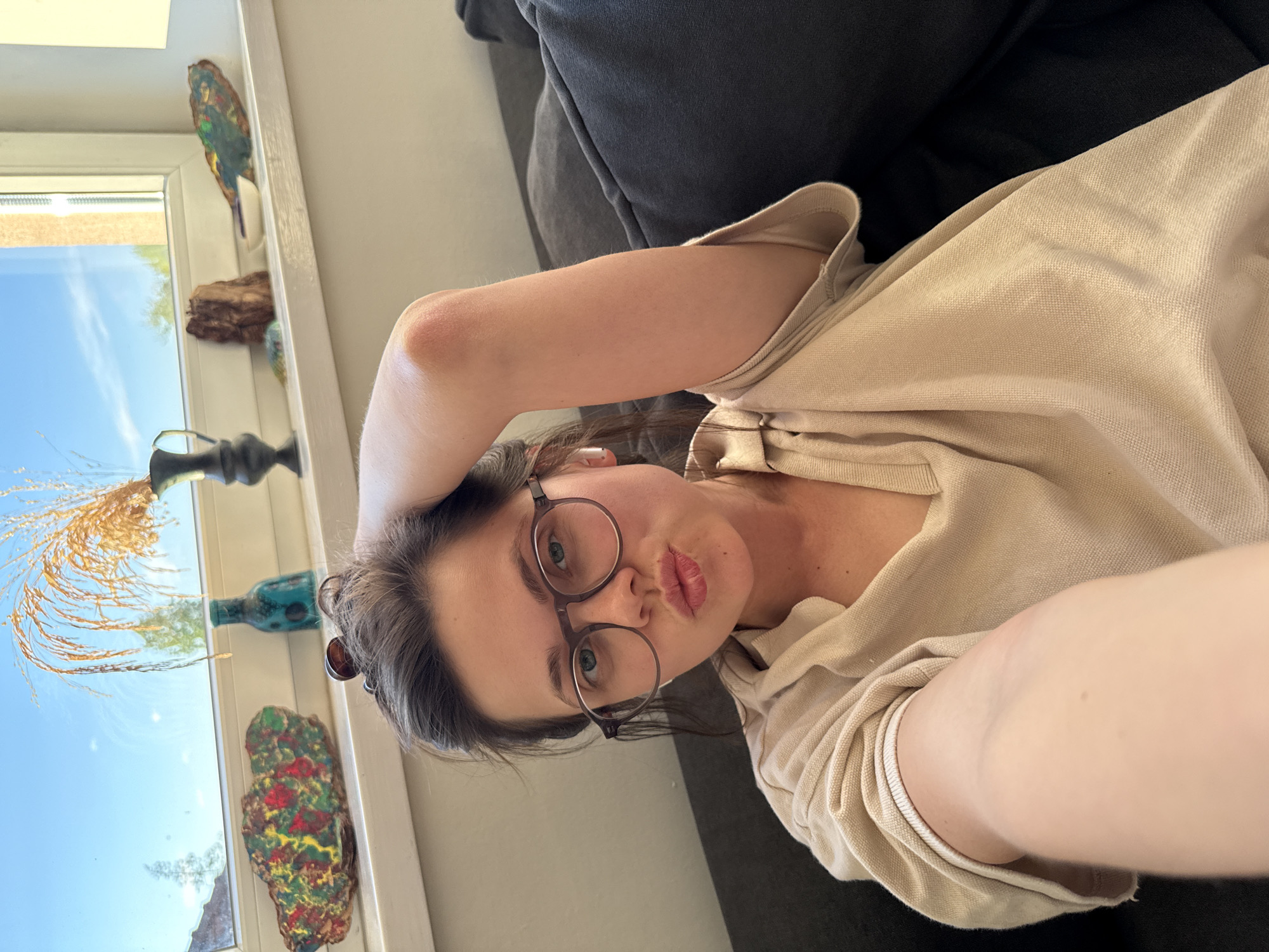 A young woman takes a selfie while reclining indoors during the daytime. The image is rotated 90 degrees clockwise. She has light skin, brown hair pulled back, and is wearing round-framed glasses. Her lips are puckered in a playful "duck face" or kissy expression, and she looks directly at the camera. She is dressed in a light beige or cream-colored hooded top, possibly a hoodie or polo shirt, with a ribbed cuff visible on her extended arm. Her arm is bent, possibly supporting her head or positioned for the selfie.
She appears to be lying on a dark grey or black couch. To her left, a window provides natural light, revealing a clear blue sky outside. The windowsill below the window is adorned with several decorative items. These include a dark, ornate vase holding dried golden grasses or reeds, a teal or blue glass bottle, a colorful, irregularly shaped piece of rock or sculpture with red, green, and yellow patterns, and a carved or painted bird figurine with blue, green, and yellow plumage perched on the window frame. There are no visible texts or specific outdoor features that indicate the location as Budapest, Hungary, within the image itself. The overall mood is casual and informal.