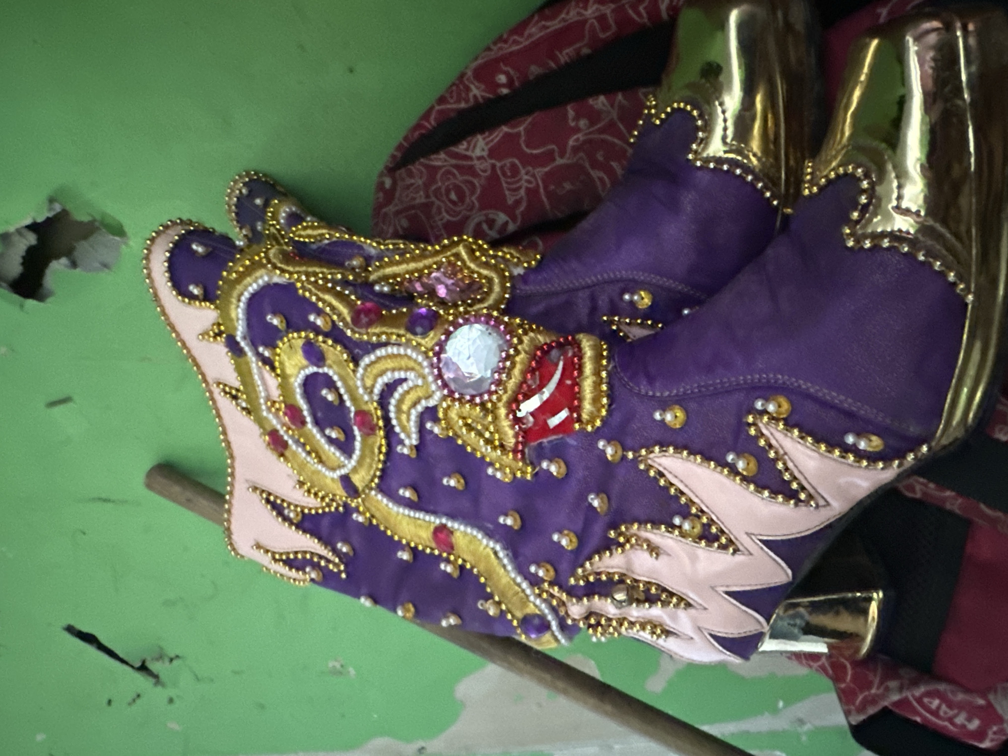 A pair of purple and gold ornamental boots lies on a surface in front of a green wall, likely in a room in Arica, Chile. The boots feature elaborate beaded designs, with a large central decorative piece on the boot that's closer to the viewer. The wall is cracked and shows signs of damage. A dark red patterned cloth rests behind the boots. A thin, light brown stick lies beneath the boot that's further from the viewer.