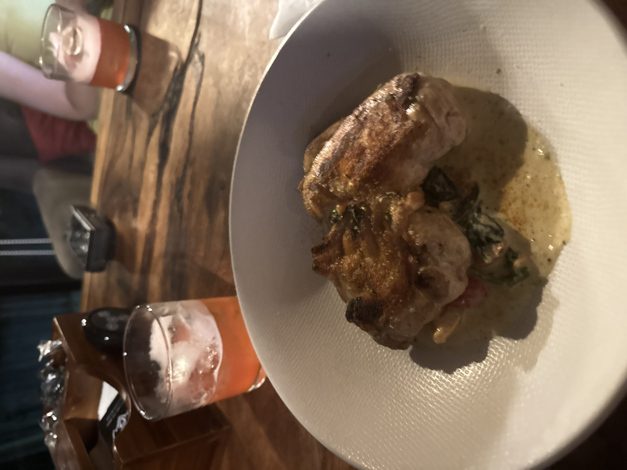 The image depicts a plate of food featuring two pieces of roasted meat accompanied by a creamy sauce and vegetables. Adjacent to the plate, there are two glasses containing beverages, one with ice. The setting appears to be a restaurant, with the scene situated on a wooden table. The location is Unknown, Unknown.