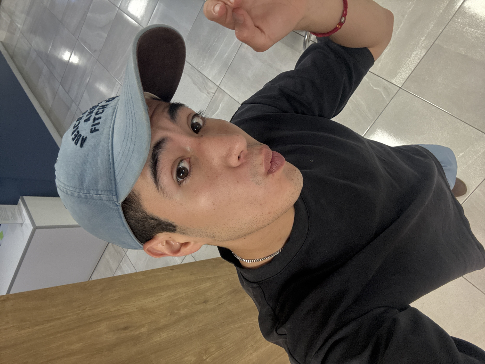 A young man is captured in a selfie taken from a high angle. He's wearing a light blue baseball cap with the word "FITCH" partially visible, a black t-shirt, and a silver chain. He is making a pouting face and extending his right arm upwards. A red bracelet is visible on his right wrist. The background consists of light-colored tile flooring and glimpses of a wooden surface in the lower left corner. The location is possibly indoors with overhead lighting. The city in Puebla, Mexico, is not explicitly identifiable from the image.