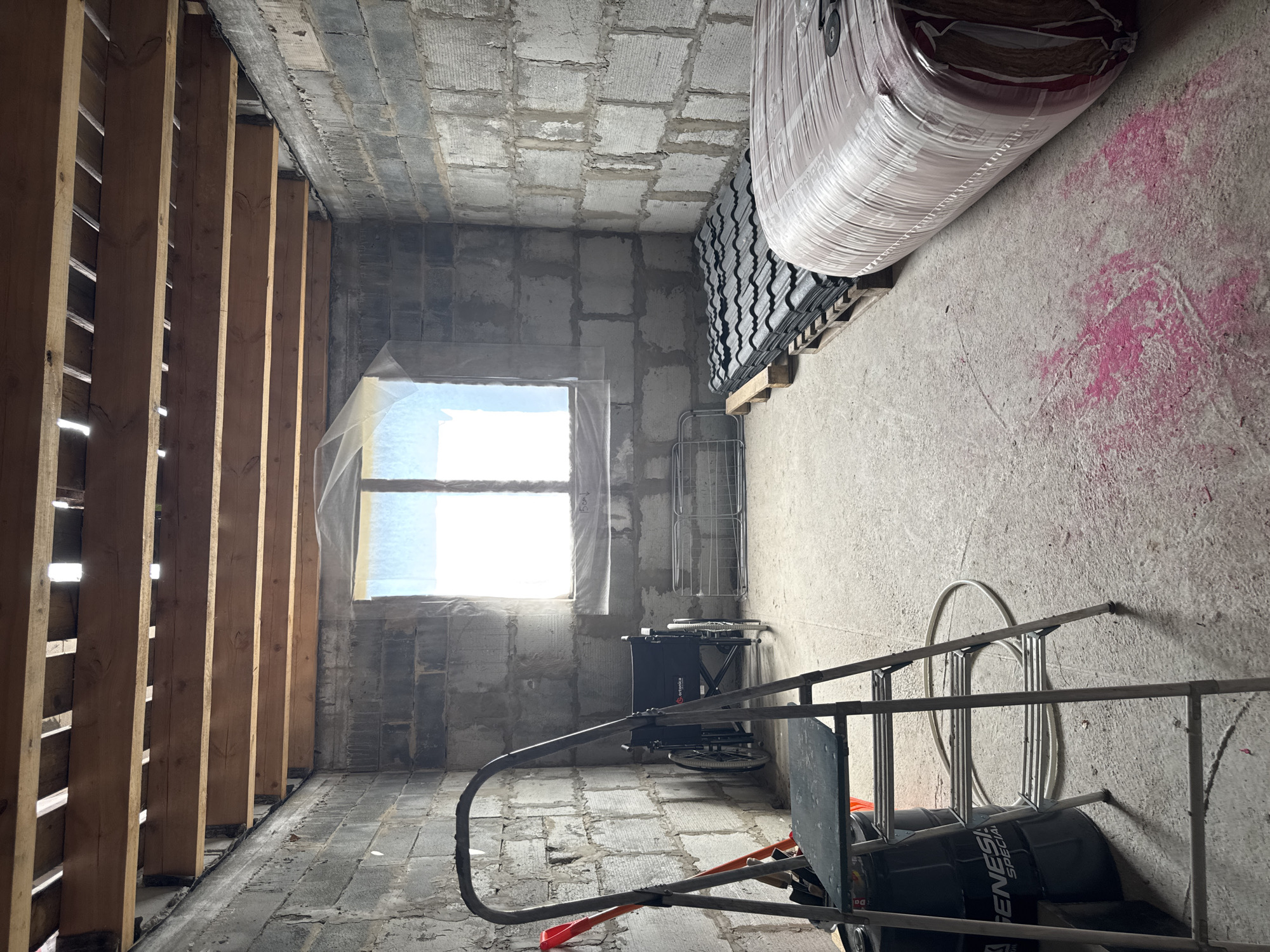 An indoor scene likely in Perm, Russia, shows an unfinished room under construction. Walls are made of concrete blocks, with a window covered in plastic sheeting. A wooden frame stands against one wall, likely for insulation or framing. Building materials are stacked on the floor, including rolls of insulation wrapped in plastic and stacks of black roofing tiles. Construction tools, including a ladder and a wheelchair, are visible, suggesting the space may be being adapted for accessibility. The floor is concrete, marked with pink paint splatters. The overall environment is one of ongoing construction and renovation.