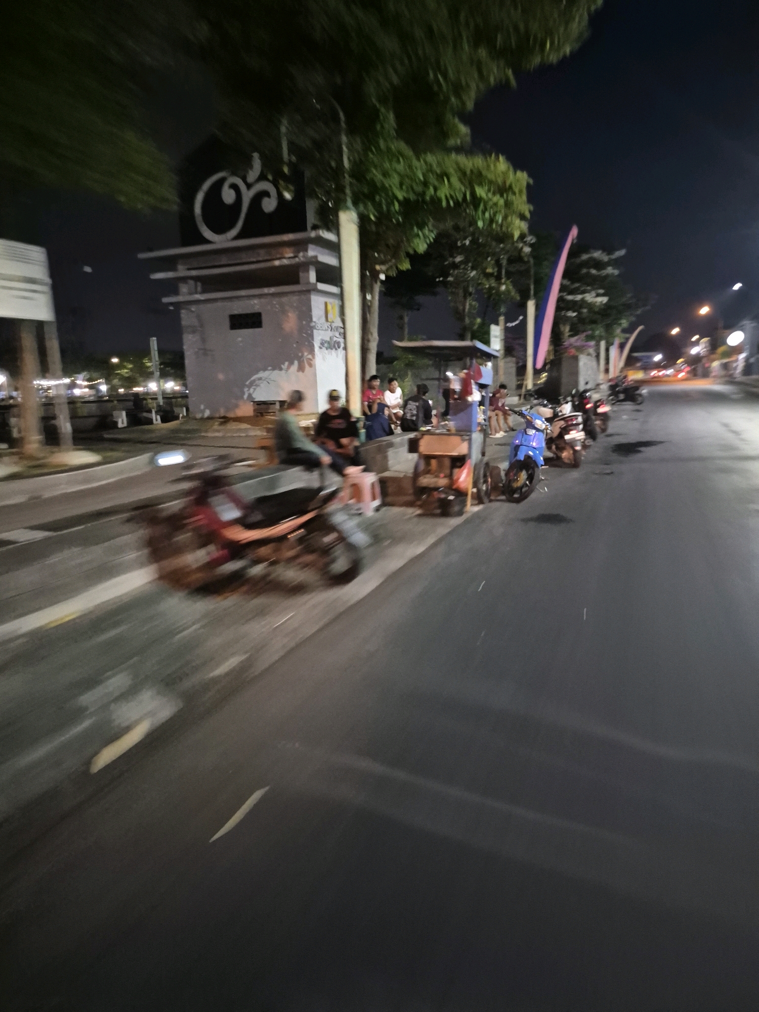 The image depicts a street scene at night in Surakarta, Indonesia. A red motorcycle is in motion on the left side of the frame, creating a motion blur. Several other motorcycles are parked along the right side of the road. A group of people are seated on stools or benches near a small structure with a decorative emblem. Streetlights and the lights from distant buildings illuminate the scene. Trees are visible along the roadside, and flags are planted near the structure. The overall impression is a casual gathering of people on a street at night, with motorcycles being a prominent mode of transportation.