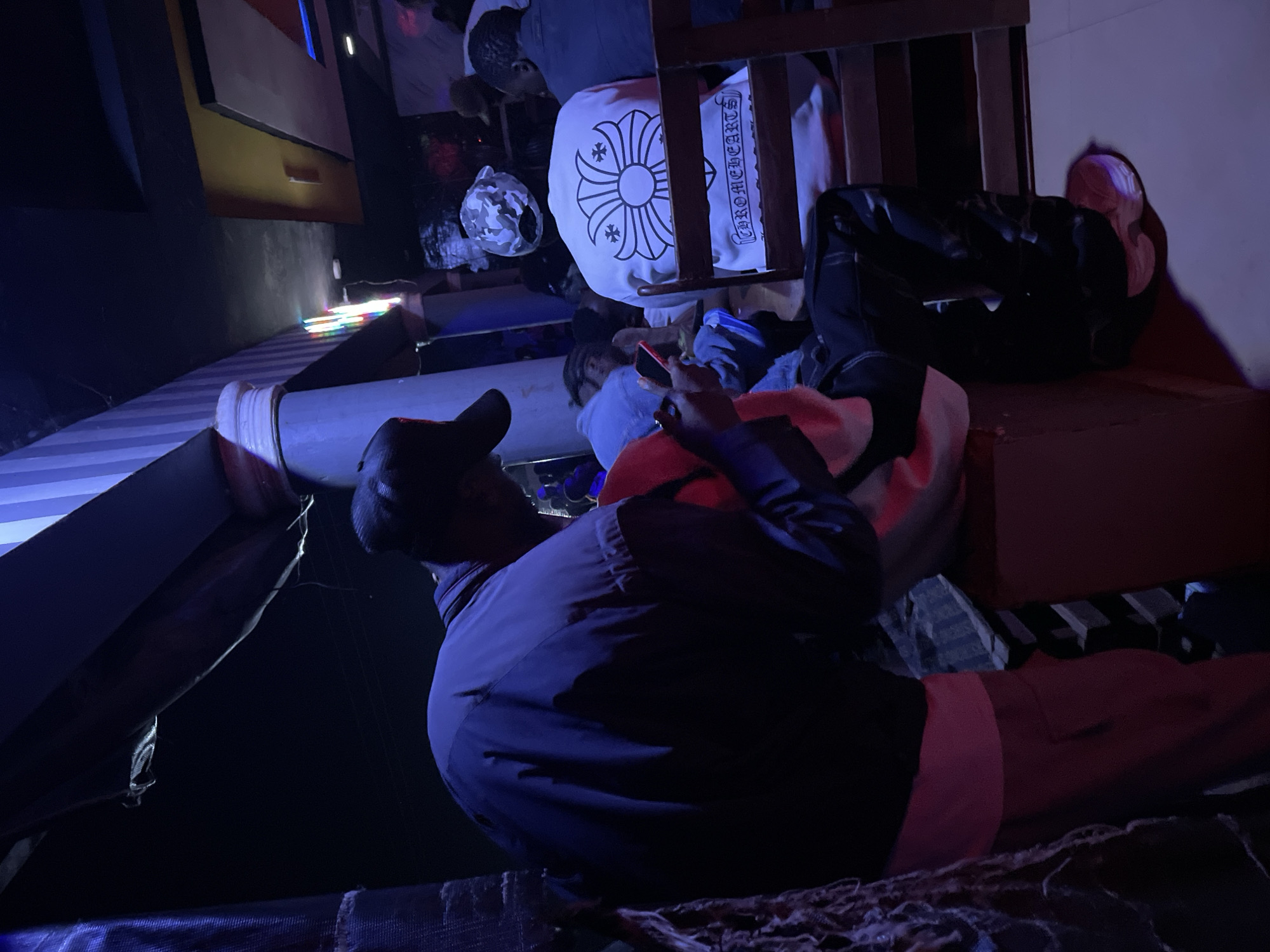 The media file depicts an indoor scene, possibly a dimly lit gathering or party, with several individuals present. The lighting is predominantly purple and blue, suggesting it's nighttime or a club-like atmosphere. In the foreground, a person wearing a dark blue jacket and a black baseball cap is looking down at their phone. Their body is contorted as they lean against a surface.
Behind this person, other individuals are visible, some lying down or seated. One person is wearing a white hoodie with a black floral design and the text "CHROMEHEARTS" in black lettering. Another person appears to be wearing black pants. The scene seems to be set on tiered platforms or a stage-like structure, with striped fabric and what looks like a large white cylindrical object in the background. Colorful, thin lights are also visible in the background, adding to the vibrant, albeit subdued, ambiance. The overall impression is one of a casual, relaxed gathering in a space with distinct, possibly artistic or themed, decor. The time of day is likely evening or night, given the artificial lighting. The emotions conveyed are neutral, with individuals seemingly engaged in their own activities or resting. There are no direct cues about the weather, as the scene is entirely indoors. The location context provided is Durumi, Nigeria, but there are no specific visual elements within the image that definitively point to this location.
