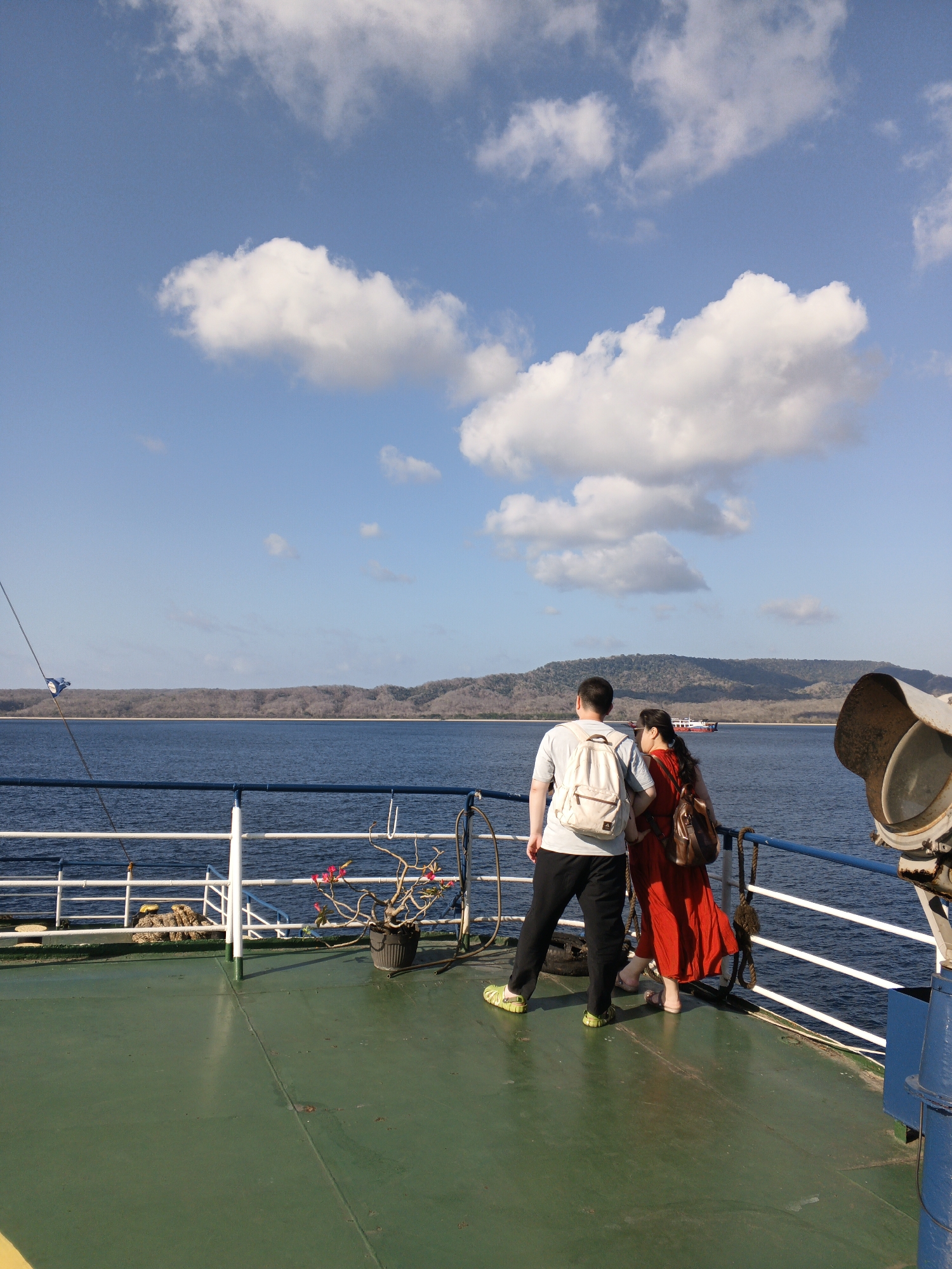 A man and a woman stand on the deck of a ferry, looking out at a body of water and a distant, tree-covered island under a bright blue sky with scattered white clouds. The man, facing away from the camera, wears a light-colored t-shirt, black pants, and lime green clogs. He has a white backpack slung over his left shoulder. The woman, also facing away, is in a vibrant red dress and tan sandals, carrying a brown backpack over her right shoulder.
The ferry deck is painted a faded green, with a white railing separating it from the water. To the left, a white rope is secured to a blue fixture. A potted plant with pink flowers sits near the railing. In the background, the water is a deep blue, and the island coastline is lined with trees. A red and white boat is visible on the horizon. To the right of the frame, a large, weathered metal light fixture is mounted. The scene suggests a ferry crossing in Indonesia, possibly between islands or to a coastal town.