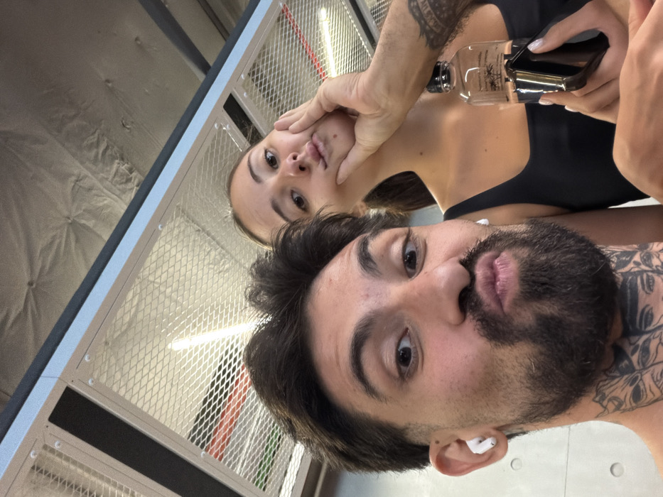 A selfie-style photograph depicts two individuals posing indoors, likely within a modern gym or recreational facility in Bangkok, Thailand.
In the foreground, an adult male with dark hair, a full beard, and a mustache is visible. He wears white wireless earbuds in both ears and has prominent tattoos on his neck depicting stylized faces. He is looking directly at the camera with wide eyes and puckered lips in a "duck face" expression.
Behind him and to the right, an adult female with dark hair, pulled back, is visible. She wears a dark square-neck top. She also exhibits a "duck face" expression, with her left hand lightly cupping her left cheek. Her left hand holds a clear, rectangular plastic bottle with a black cap and dark liquid, identifiable by a label featuring a palm tree logo and text including "Pure Coco," "Chaweng," and "Koh Samui."
The man's right arm extends towards the woman, with his hand resting on her lower left cheek.
The background features an industrial-style ceiling with visible white expanded metal mesh panels, exposed structural elements, and a vertical fluorescent light fixture. A light grey wall with small circular indentations is partially visible on the far left.