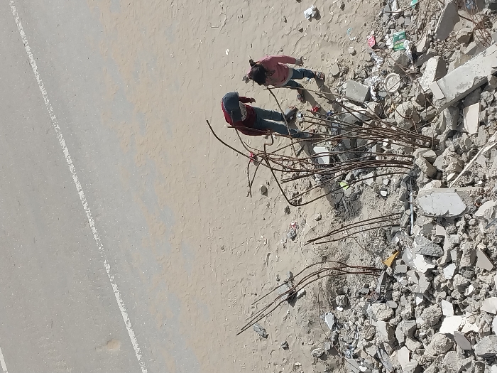 The image presents a high-angle, almost aerial view of an outdoor scene featuring two individuals amidst what appears to be a demolition or construction site. The scene is divided roughly into two main areas: a relatively clear ground expanse and an area filled with rubble and debris.
On the left side of the frame, there's a light gray-beige asphalt road, marked by a faded white dashed line running vertically. Adjacent to the road, extending towards the right, is a wider stretch of light-colored, sandy or dusty ground. This ground appears somewhat uneven and scattered with small, faint irregularities.
To the right, the ground transitions into a large pile of varied debris. This rubble consists primarily of broken concrete pieces, varying in size from small fragments to larger chunks. Numerous twisted and bent metal reinforcing bars (rebar) protrude from and are entangled within the debris, suggesting the remnants of a concrete structure. Scattered among the rubble are also a few pieces of what looks like plastic or discarded packaging.
Two individuals are visible on the sandy ground, positioned near the edge of the debris pile. The person closer to the road (and further left) is wearing a red long-sleeved shirt and dark pants. This individual is bent over, facing towards the debris, and appears to be focused on something on the ground or within the lower part of the rubble. The second person, positioned slightly to the right and closer to the main pile of debris, is wearing a lighter-colored (possibly pink or light red) long-sleeved shirt and blue jeans. This individual is standing more upright, looking towards the debris.
The lighting indicates a bright, sunny daytime, casting clear but subtle shadows. The overall impression is that of manual labor or inspection occurring at a site of structural dismantling, damage, or reconstruction. No specific text or distinct geographical markers are visible in the image.