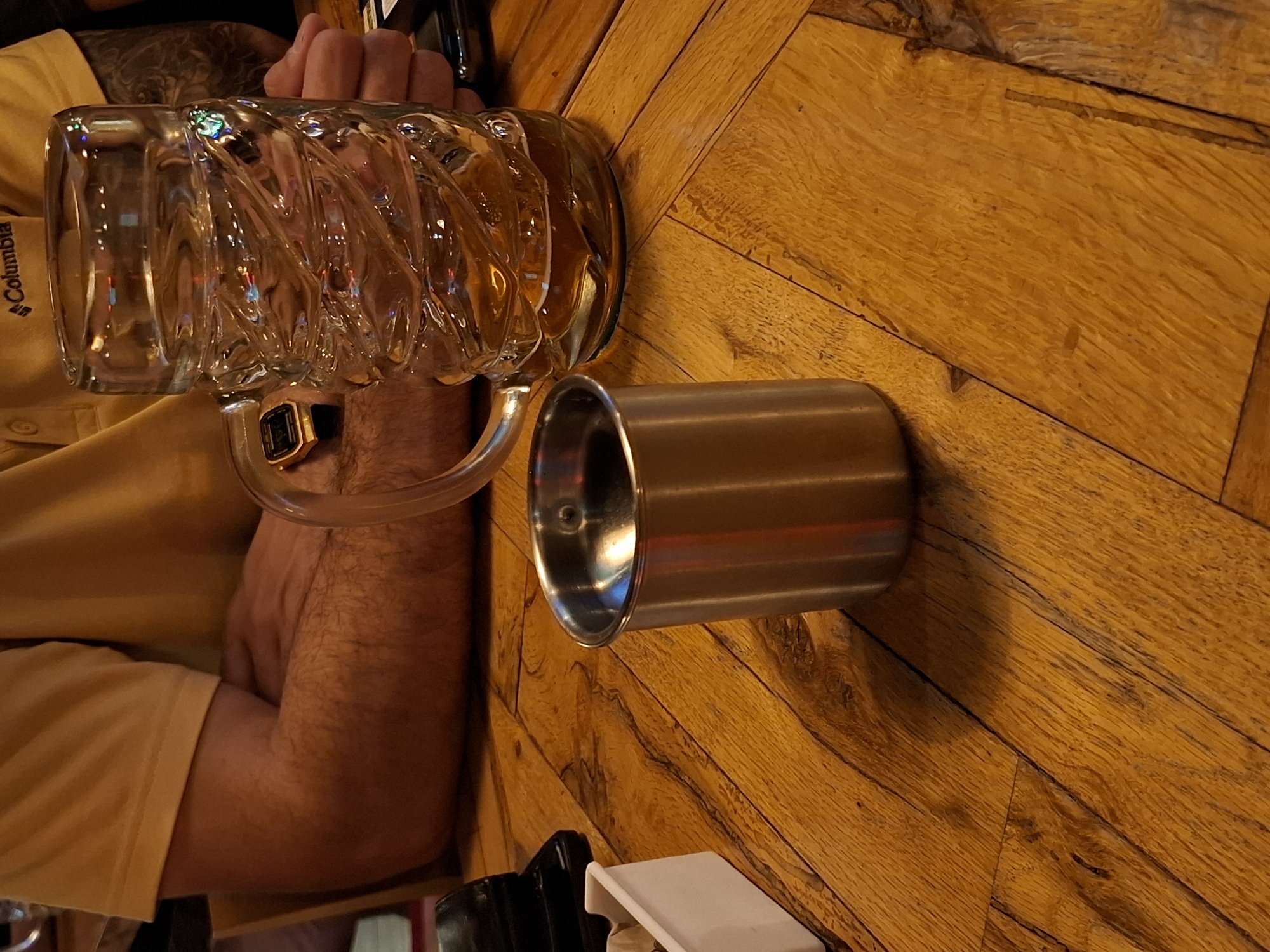 In an indoor setting with a warm, woody ambiance, a person is seated at a table. The individual, whose arms are visible, is wearing a yellow t-shirt with a Columbia logo. A large, textured glass mug filled with a amber liquid sits on the table to the right, its handle to the left. To the left of the mug, a small, metallic cup with a brushed finish is positioned. In the background, a white rectangular container with something light-colored inside is partially visible on the left. The table surface, made of wooden planks, exhibits a prominent grain pattern. The lighting casts shadows, indicating a light source from the upper left.