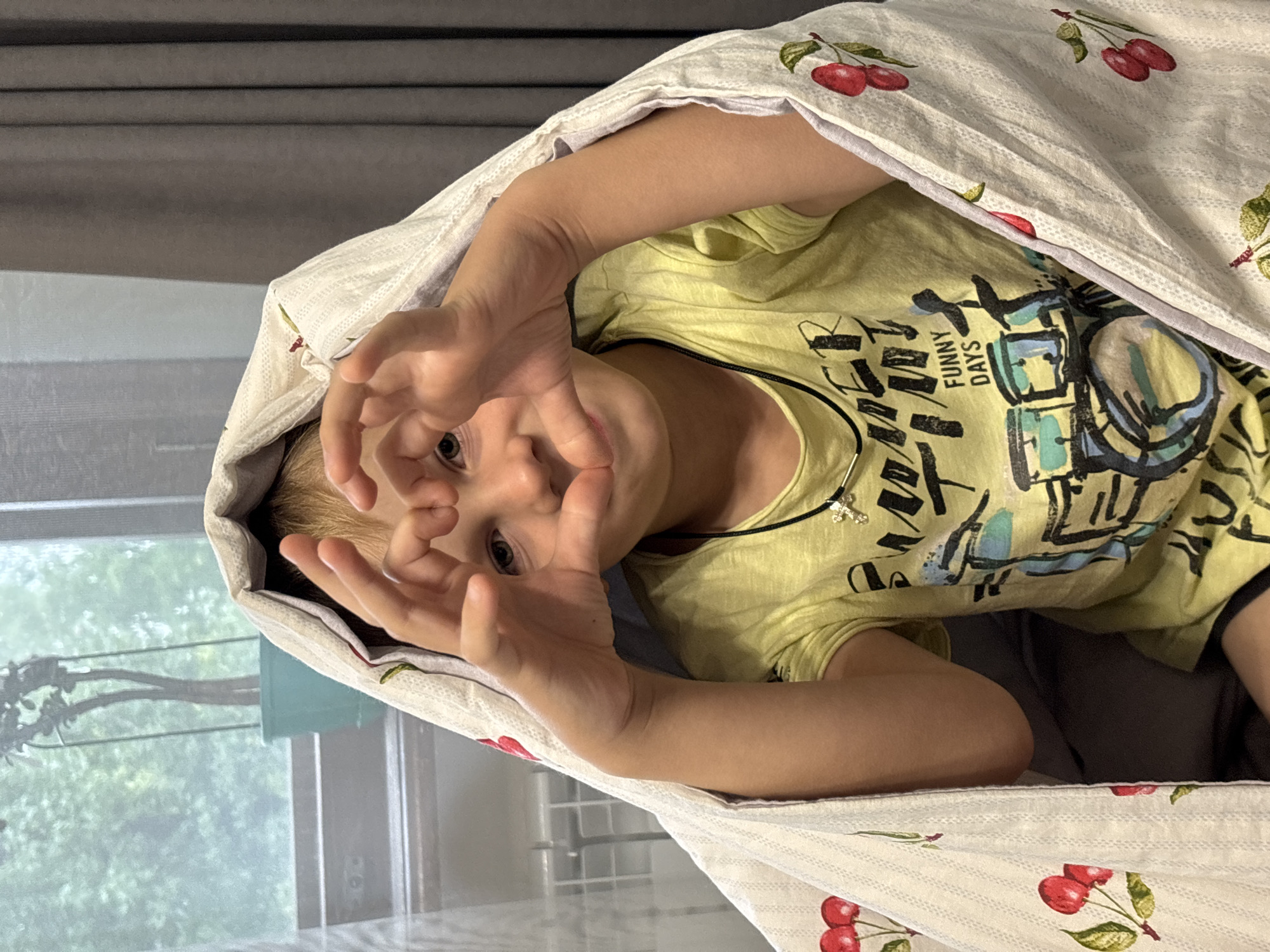 A young child, with blonde hair and light skin, is peeking out from under a white blanket with a cherry pattern. The child is wearing a yellow t-shirt with a graphic print and a black necklace with a cross pendant. The child is holding their hands up to their face, forming a heart shape. The background shows a window with a plant and a grey curtain. The scene appears to be indoors, possibly in a bedroom, with a radiator visible on the left. The location is identified as Rusanovo, Russia.