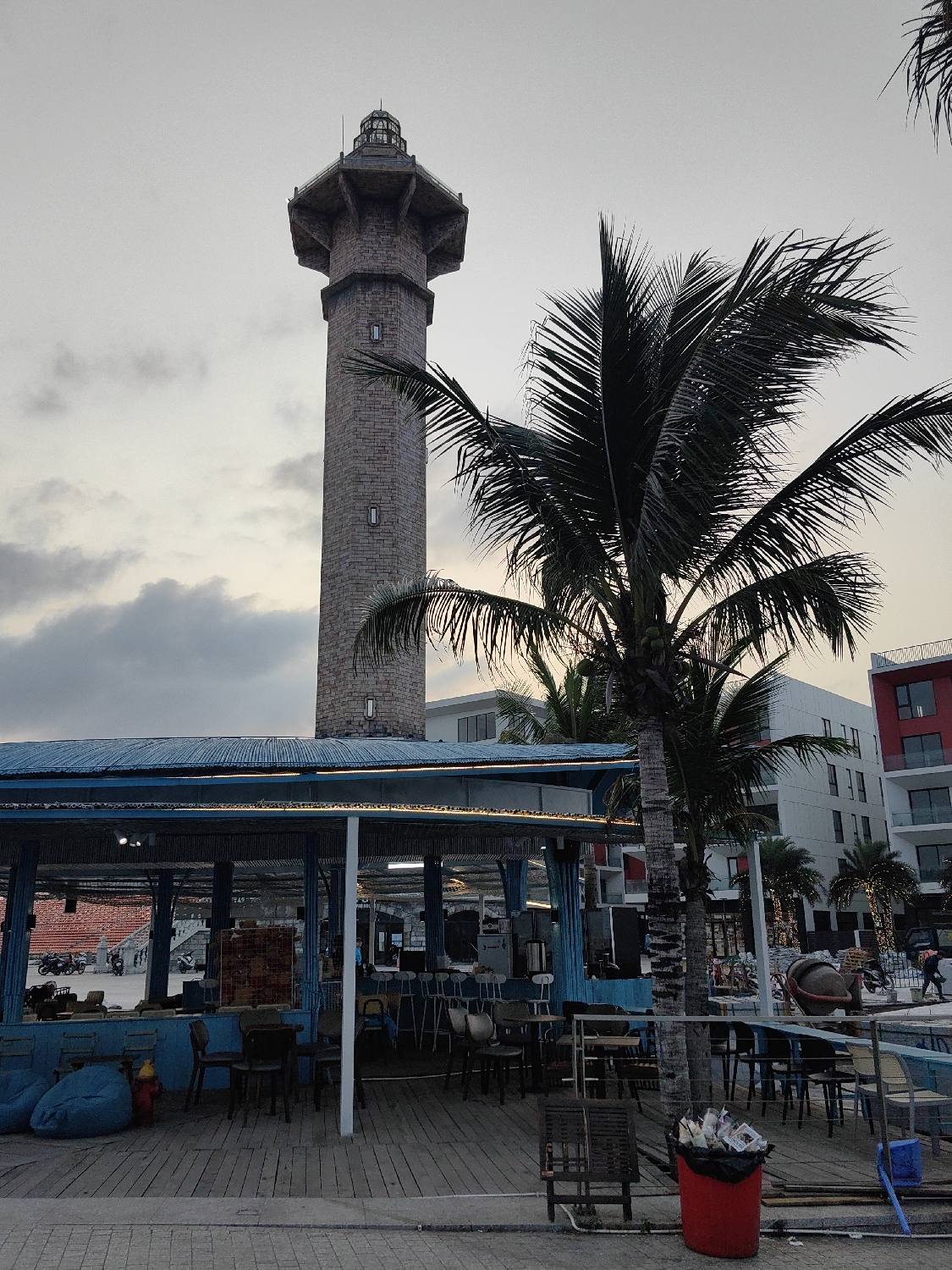 The image captures a dusk scene in Bai Chay Ward, Vietnam, featuring a tall, stone lighthouse, an outdoor bar, and palm trees under an overcast sky.
In the left half of the frame, a prominent, slender lighthouse stands tall, constructed from light-colored stone blocks. Its base is cylindrical, transitioning to an octagonal section near the top, which culminates in a glass-enclosed lantern room with a conical cap. Small, rectangular windows are visible along its shaft. The sky behind the lighthouse is a muted gray, suggesting either late afternoon or early evening.
In the foreground and central to the image is an open-air bar or restaurant structure. It features a corrugated blue roof supported by blue metal posts, with string lights illuminating the underside of the canopy. The establishment is set on a wooden deck, furnished with various tables, chairs, and several blue bean bags. A bar counter with stools is visible inside. To the left, a red fire hydrant is positioned near the bean bags.
A large palm tree with lush, dark green fronds stands in front of the bar, its trunk partially obscured by the bar's structure, and its foliage reaching towards the lighthouse, partially overlapping it. Another palm tree is visible further to the right.
In the background, beyond the bar and palm trees, are modern multi-story buildings, some with distinctive red and white facade elements. Construction materials, including a large concrete mixer, are visible on the right, suggesting ongoing development. A few indistinct figures can be seen in the distance, near the buildings and inside the bar area, but no specific activity is clearly discernible. A red trash can, overflowing with what appears to be discarded paper or plastic, sits on the wooden deck in the lower right.
The overall atmosphere is casual and relaxed, indicative of a coastal entertainment area during the transition from day to night. There is no clearly legible text visible in the image.