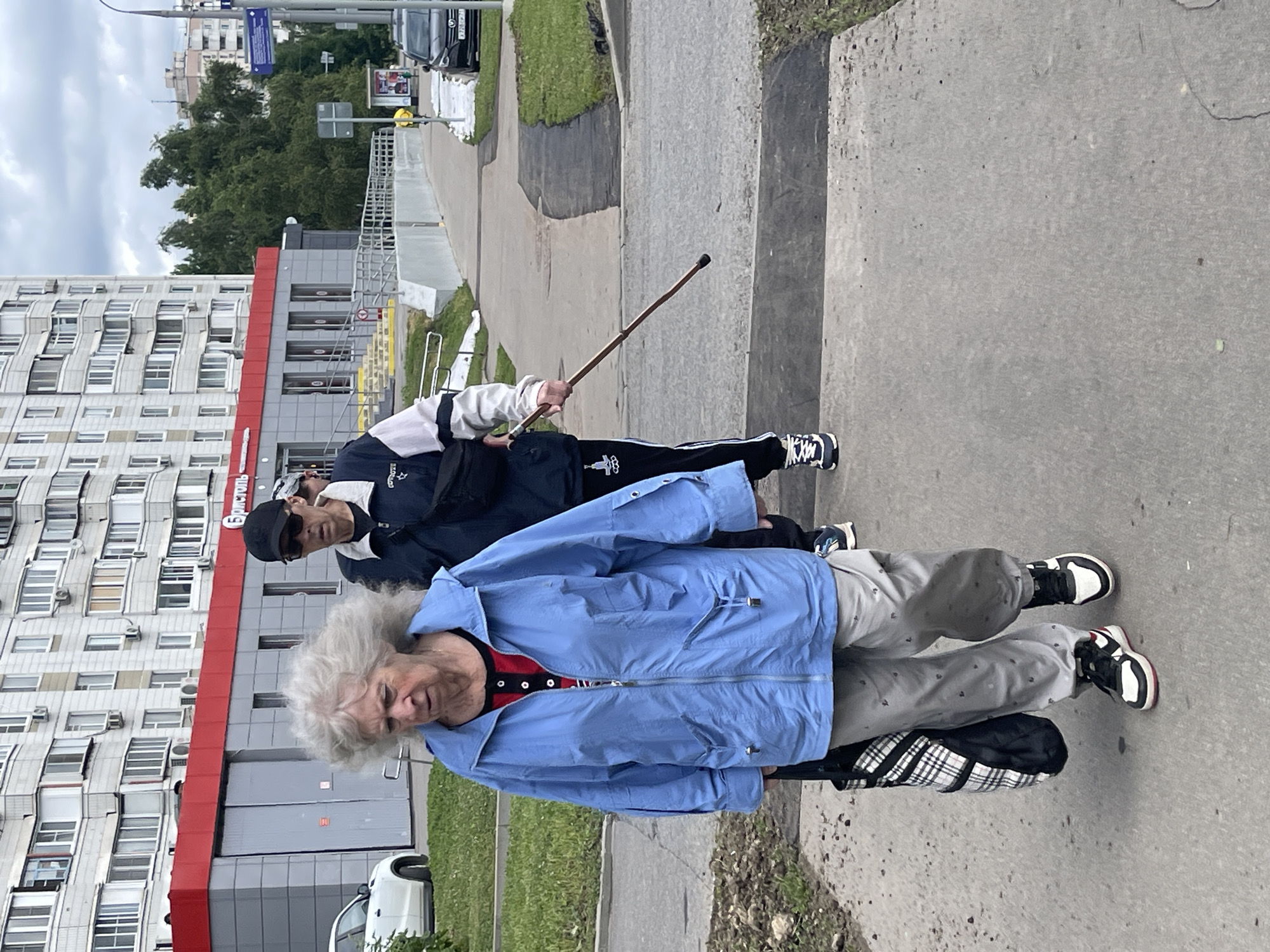 A person in a blue jacket and grey pants walks on a sidewalk in Moscow, Russia. The person is carrying a black and white plaid bag in their left hand. Another person in a dark jacket and black pants walks behind them, holding a cane. They are walking in front of a building with a red stripe on its facade. The sky is cloudy.