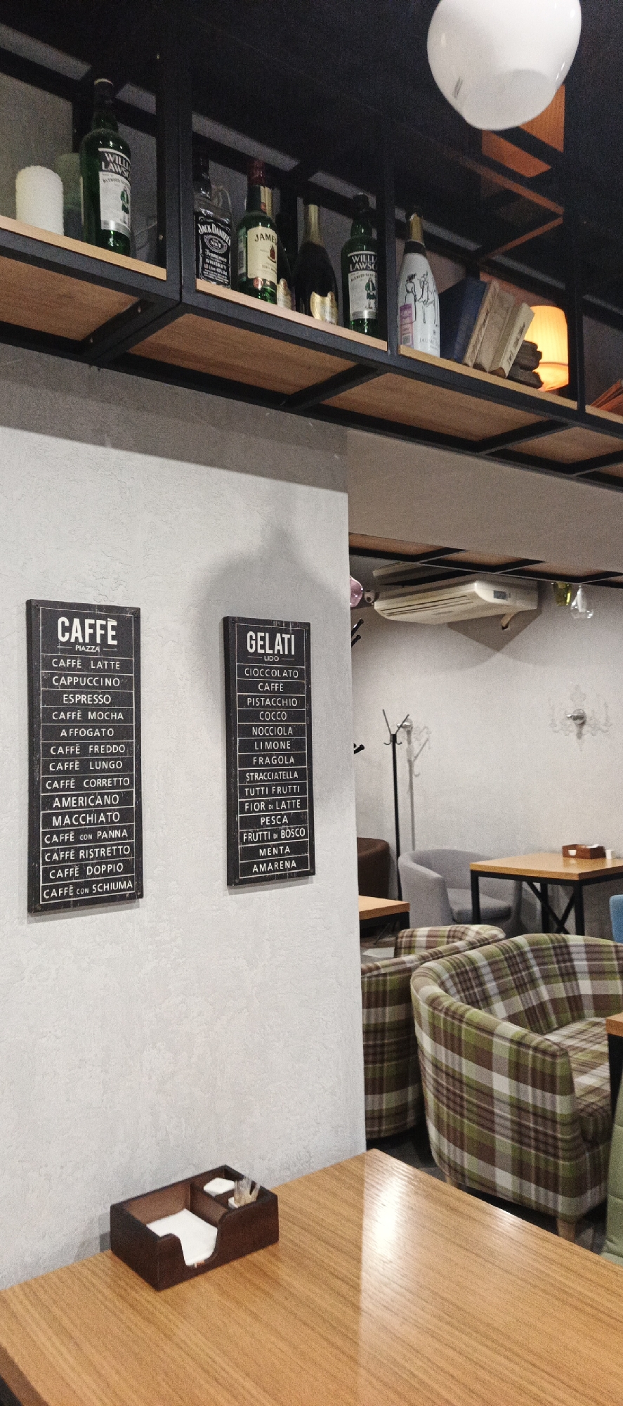 This image depicts the interior of a cafe or bar. On a shelf above, various bottles, including Jack Daniel's and Jameson whiskey, as well as other liquor and wine bottles, are visible. Below the shelf, on the wall, hang two signs: one listing "CAFFE" options such as latte, cappuccino, and espresso, and another listing "GELATI" (ice cream) flavors like cioccolato, caffè, and pistacchio. In the foreground, a wooden table holds a dark brown wooden organizer with napkins and toothpicks. Behind the table, several comfortable armchairs with plaid upholstery are arranged around other wooden tables. The lighting appears to be warm and ambient, with a large white light fixture visible at the top of the frame. The overall impression is of a cozy and relaxed establishment. The image is likely taken in Votkinsk, Russia, as indicated by external information not present in the image itself.