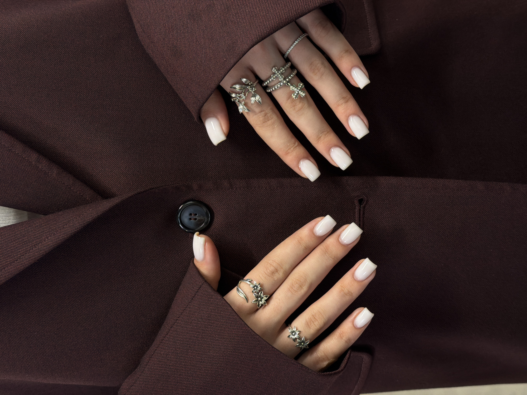 A close-up studio shot displays two hands adorned with white square-tipped nails and various silver rings. The hands are posed against a dark brown fabric, presumably a blazer or coat, featuring a single dark button. The hands are accessorized with multiple silver rings of varying designs, including floral, cross, and band styles. The background is out of focus, and the lighting appears to be artificial. The scene is reminiscent of a fashion or beauty advertisement. There are no other discernible details that would point to the city of Yerevan, Armenia.