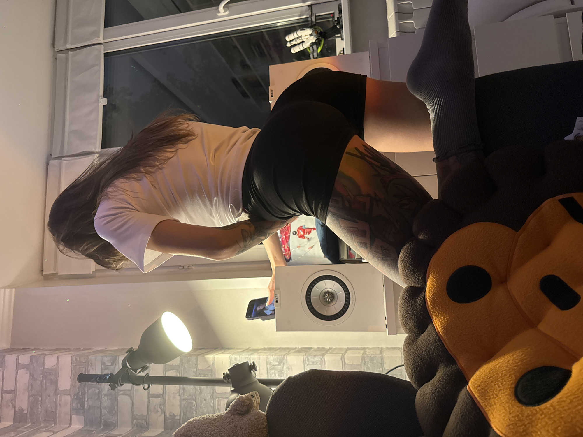 A woman is pictured indoors, possibly in an apartment, with a city view visible through a large window. The room is dimly lit, with a lamp providing illumination. The woman is wearing a white t-shirt and black shorts, and has visible tattoos on her legs. She is kneeling on a plush toy. In the background, there is a speaker, a mobile phone, and what appears to be a 3D printer. The window shows a dark exterior, suggesting it is nighttime. The overall impression is a candid shot within a residential setting.