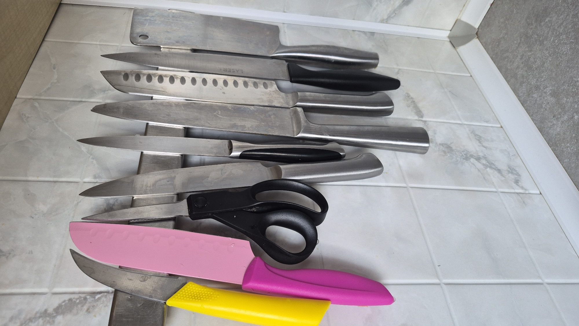 In a kitchen, likely in Krauja, Latvia, a magnetic knife strip is mounted on a tiled backsplash. Several knives and a pair of black-handled scissors are attached to the strip. From left to right, the objects visible are: a yellow-handled knife, a pink-handled knife with a broad blade, black-handled scissors, a knife with a silver handle, a knife with a silver handle and black accents, a knife with a silver handle and a series of circular indentations on its blade, a knife with a silver handle and black accents, and a large cleaver-style knife with a silver blade and handle. The backsplash tiles are a light color with a marble-like pattern. The countertop below the backsplash is a grey speckled material. The overall scene depicts organized kitchen utensils.