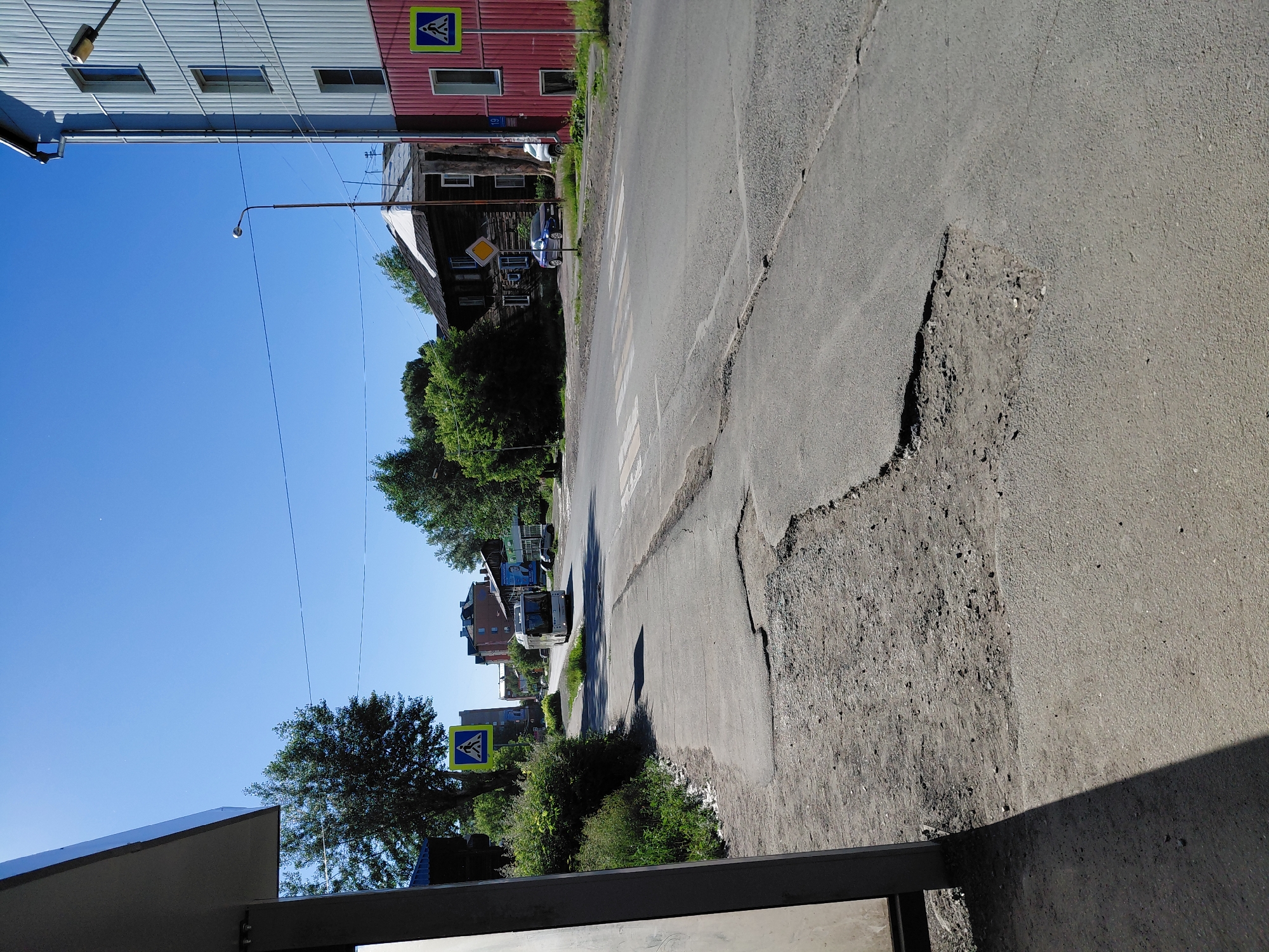 The image depicts a street scene in a city. In the foreground, a paved walkway runs alongside a road. The walkway is cracked and shows signs of disrepair, with several large potholes. To the left of the walkway is a bus stop shelter, casting a shadow. The road itself appears to be an asphalt surface with a visible crosswalk. Several vehicles are present, including a bus traveling down the road and a blue car parked on the side. Buildings line the street, ranging from multi-story structures to older wooden houses. Green trees and vegetation are scattered throughout the scene. The sky is clear and blue, indicating a sunny day. Two pedestrian crossing signs are visible, one on the left side of the road and another on the right. The overall environment suggests a somewhat urban or suburban setting with a mix of residential and commercial buildings.