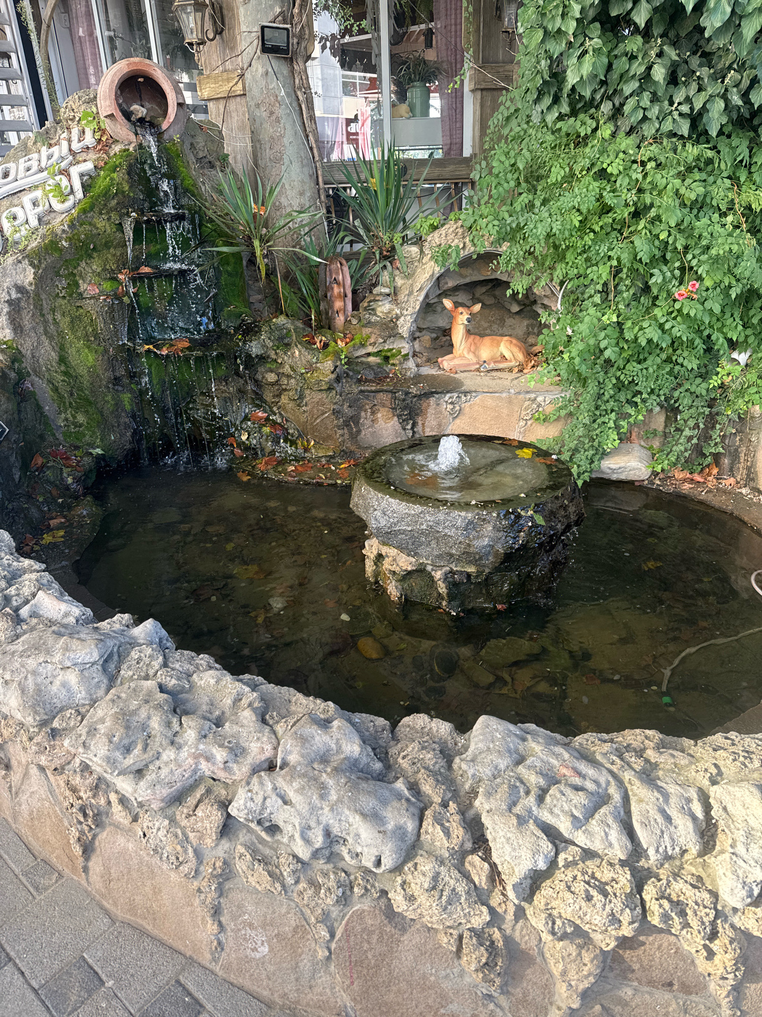 The image shows an outdoor decorative fountain in Anapa, Russia. The fountain features a tiered waterfall made of rocks and moss, with a large terracotta pot at the top from which water flows. Below the waterfall is a small pond. In an alcove to the right of the waterfall, a statue of a deer is placed amidst lush green ivy. A smaller fountain in the center of the pond shoots water upwards. The surrounding area is paved with interlocking bricks and lined with a rough stone border. A building with large windows is visible in the background. The scene is illuminated by natural daylight.