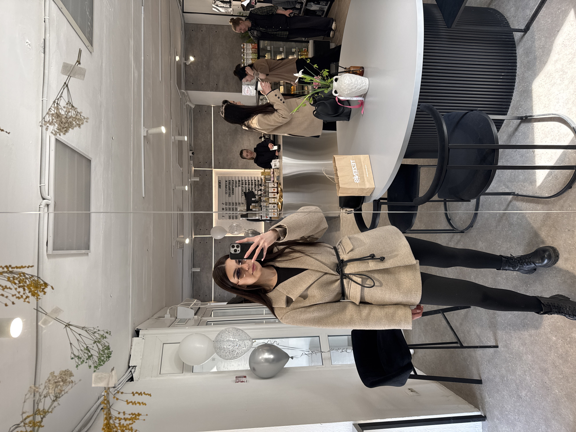 The image shows a woman taking a selfie in a restaurant. She is wearing a beige coat, black leggings, and black boots. She is holding a phone in her right hand. In the background, there are other people dining, a round table, and a black chair. There are balloons near the window, some wall decoration and plants on a table. A sign that reads "LESENKI" is visible. Based on the sign, the location appears to be the Lesenki restaurant in Chaikovsky, Russia.