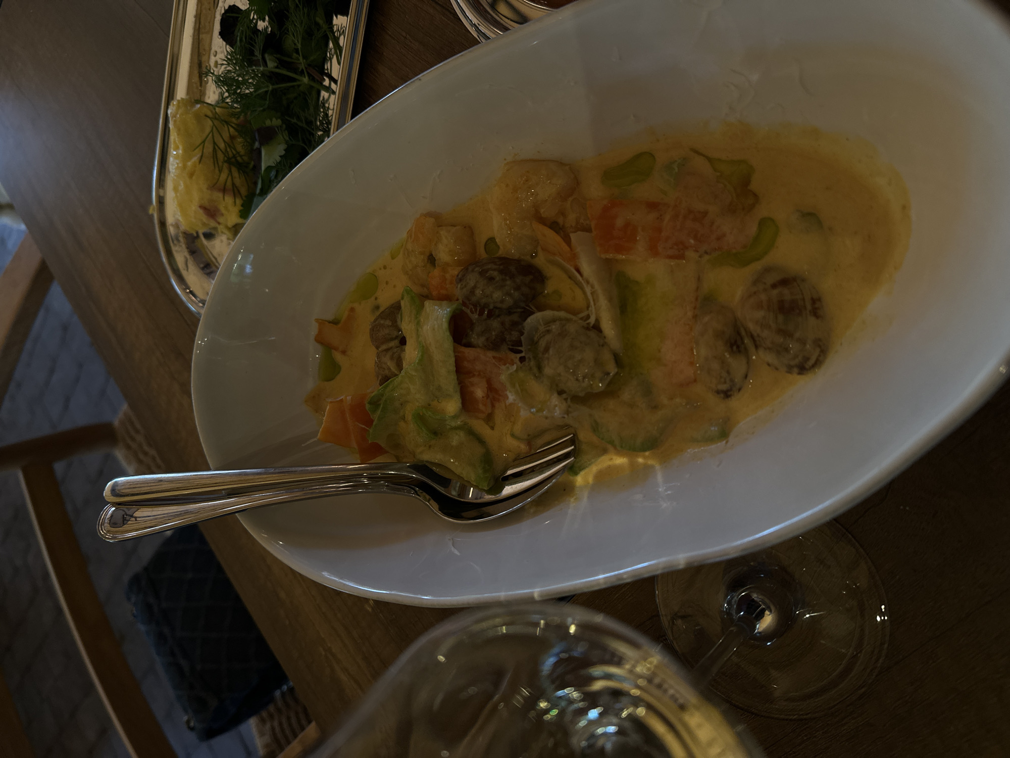 The image captures a dining scene indoors, likely in a restaurant, with the focus on a white, oval-shaped plate containing a creamy dish. The dish appears to have a light sauce and includes elements such as mushrooms, avocado, and what seems to be chopped vegetables like carrots and possibly squash. A fork rests on the plate, suggesting it is ready to be eaten.
Adjacent to the main plate is a silver dish containing a small portion of what appears to be a salad or a side dish with greens and a yellow element, which might be butter or a dressing. In the foreground, there is a wine glass partially visible, hinting at the presence of drinks alongside the meal.
The scene is set on a wooden table, contributing to a warm, possibly upscale dining atmosphere. The presence of a chair leg and the edge of a dark-colored seating cushion suggests that the photo was taken from the perspective of someone sitting at the table. The overall lighting is soft, indicating it might be evening or that the indoor lighting is creating a subdued ambiance.
Without explicit visual cues, definitively stating the location as Yerevan, Armenia, is impossible, so this detail should be disregarded.