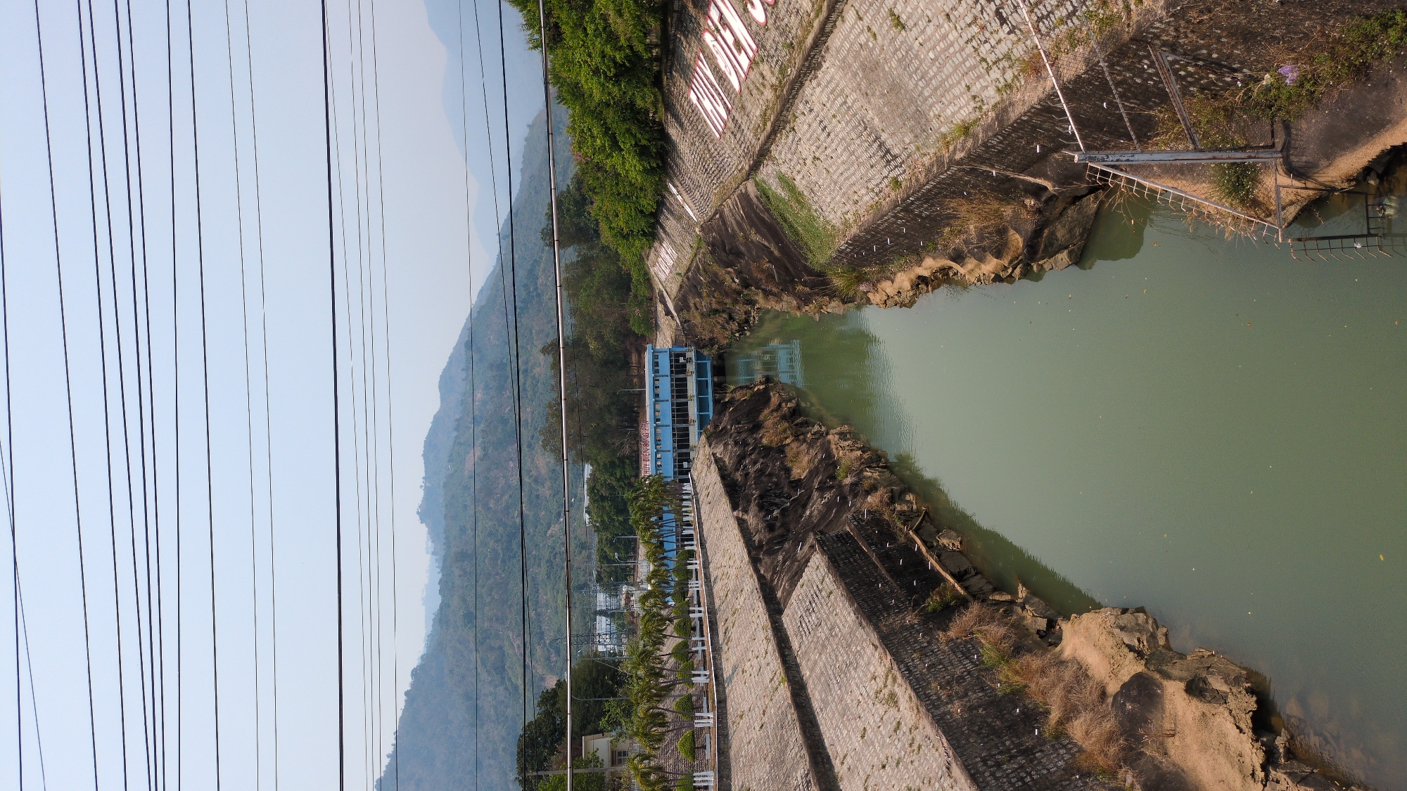 This aerial-perspective photograph shows the Dinh Dinh dam and the surrounding landscape in Lâm Sơn, Vietnam. The dam is constructed of grey stone blocks and concrete. A blue building sits near the water's edge. The water is a murky greenish-brown. In the distance, a mountain range is visible under a hazy sky. Power lines crisscross the left side of the image. The camera appears to be positioned above the water level, offering a view down into the dam and the surrounding terrain.