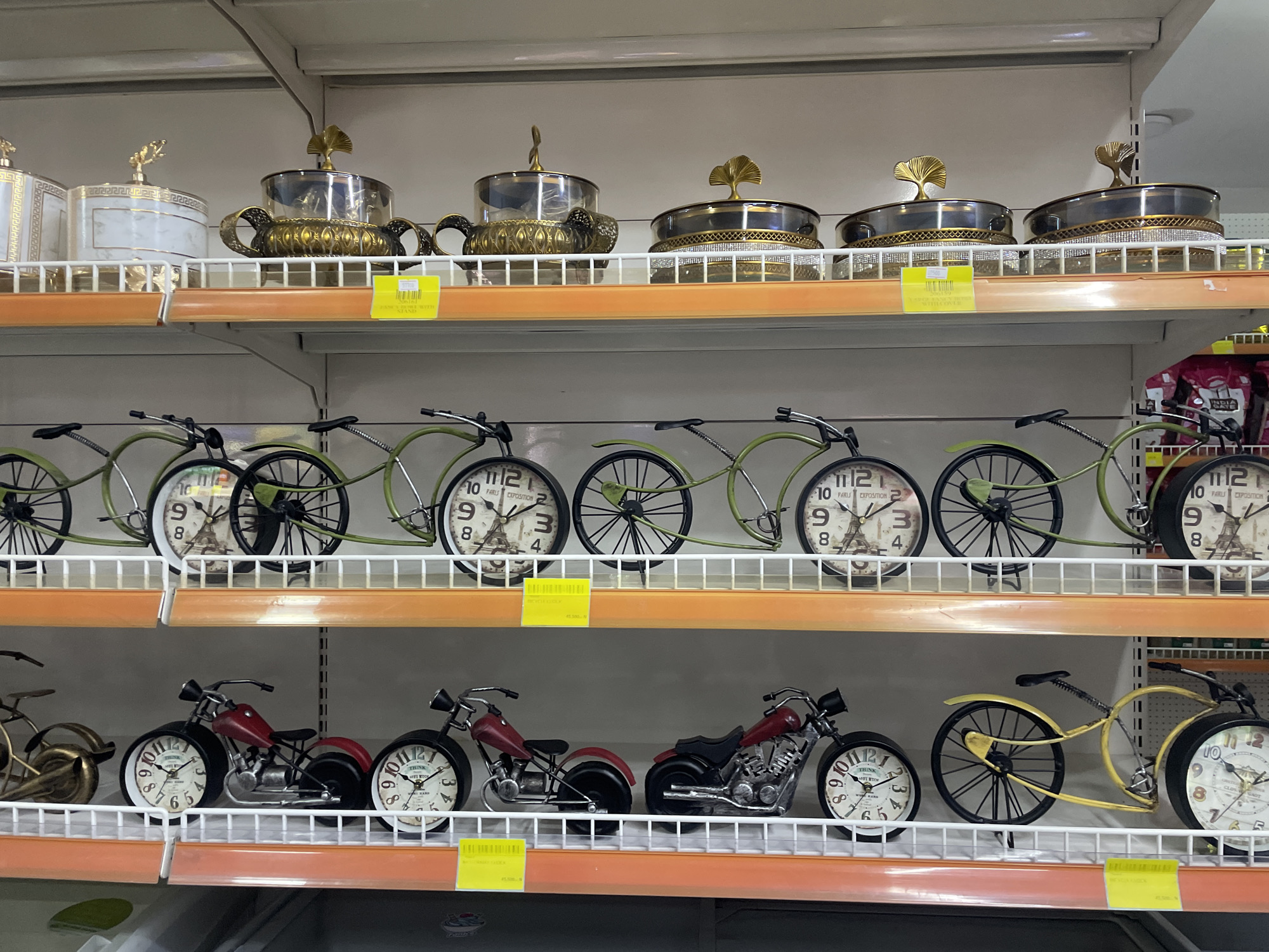 The image displays a retail store shelf in Galadimawa Village, Nigeria, showcasing various decorative items. The top shelf features ornate, lidded containers, possibly for serving or storage, with intricate gold detailing and translucent lids. Below this, the middle shelf holds several metal bicycle-shaped clocks, each with a circular clock face displaying Roman numerals and imagery. The bottom shelf exhibits metal motorcycle-shaped clocks, also featuring circular clock faces with Roman numerals, alongside a decorative yellow bicycle clock. The items are neatly arranged on white wire shelves, illuminated by overhead lighting, creating a well-lit display. The background appears to be the interior of a store, with gray shelving units and other products partially visible.
