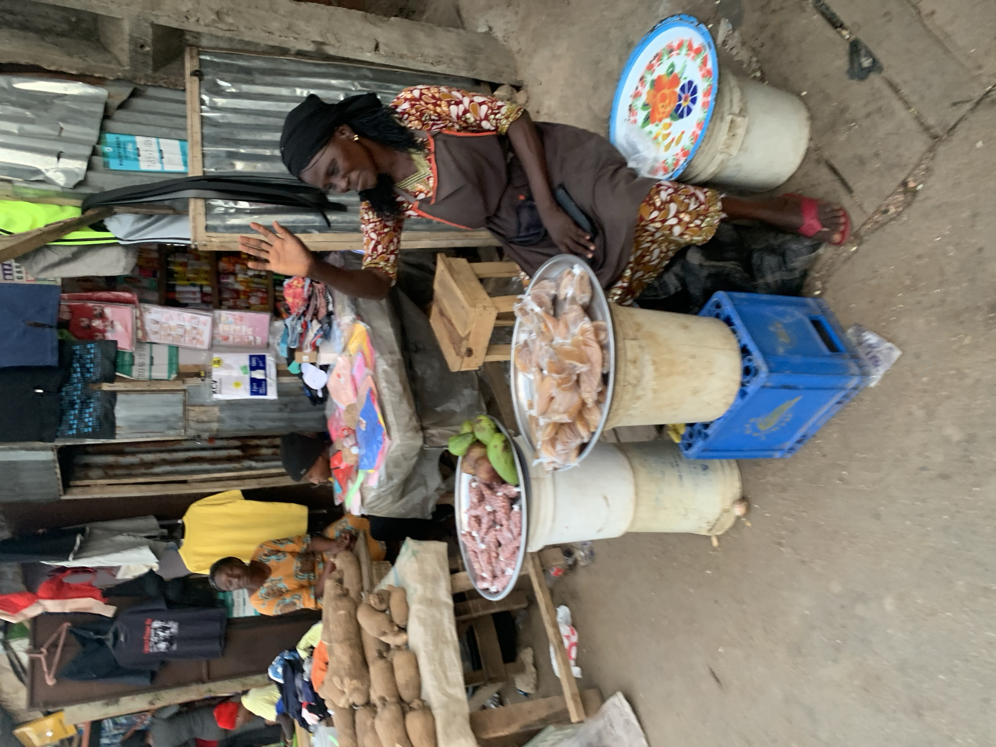 In Jos, Nigeria, an outdoor market scene is captured from a high-angle perspective. The focus is on two women selling goods at their stalls. The woman closer to the camera wears a dark head covering, a patterned blouse, and a dark apron. She is seated and gestures with her right hand. Her stall displays trays filled with wrapped food items and raw produce on top of buckets. Another woman, wearing an orange patterned shirt and yellow shirt, sits at a stall with sweet potatoes. The stalls are located next to a building with corrugated metal siding. Clothes hang for sale. The ground is concrete.