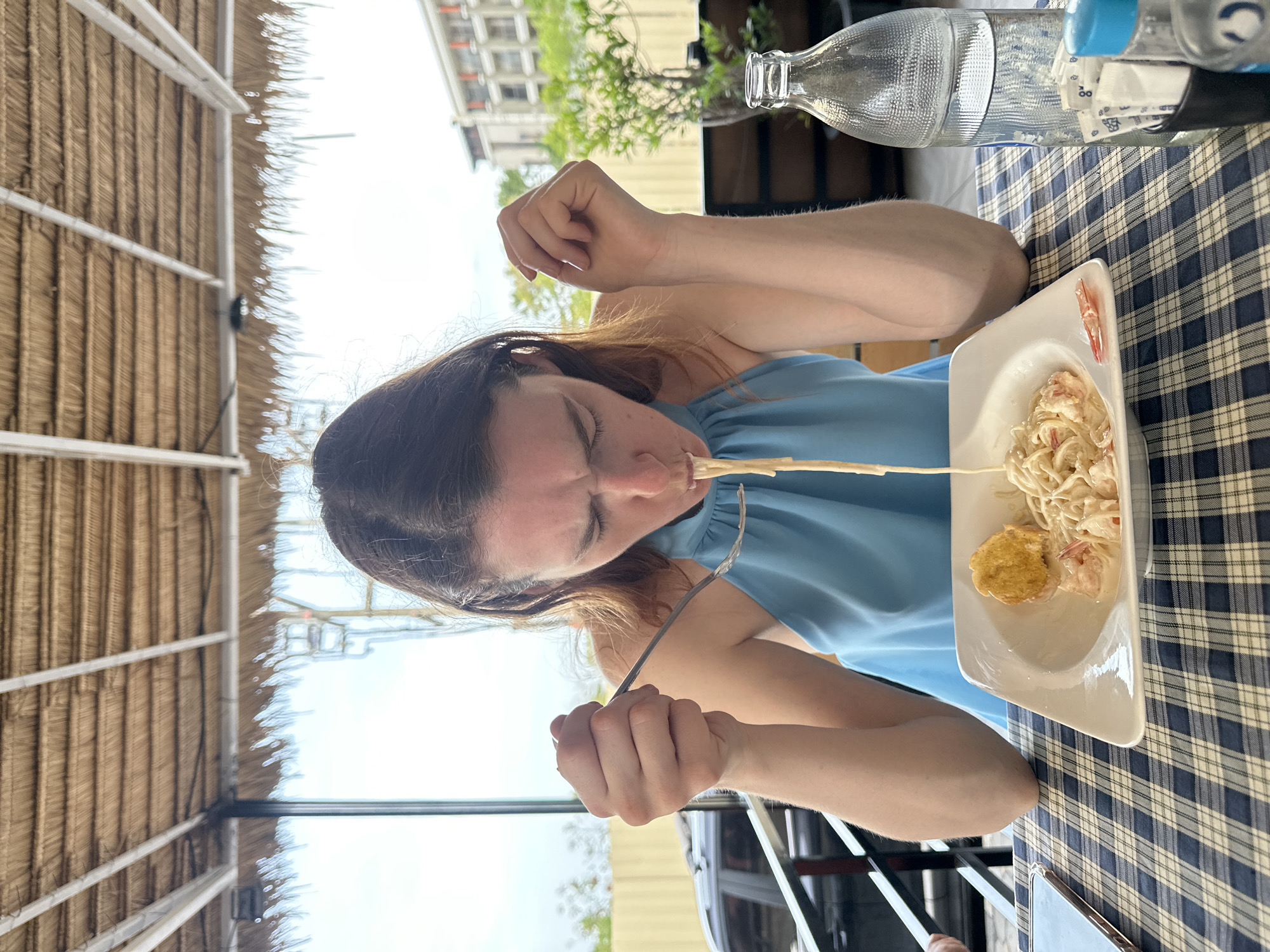 A woman is seated outdoors at a table covered with a blue and white checkered tablecloth, eating a plate of pasta with what appears to be shrimp and a side of what might be a small piece of bread or fried food. She is wearing a light blue top. In the background, a thatched roof and an urban scene including a building and greenery are visible. Two water bottles are also present on the table. The location is Baan Chaweng, Thailand.