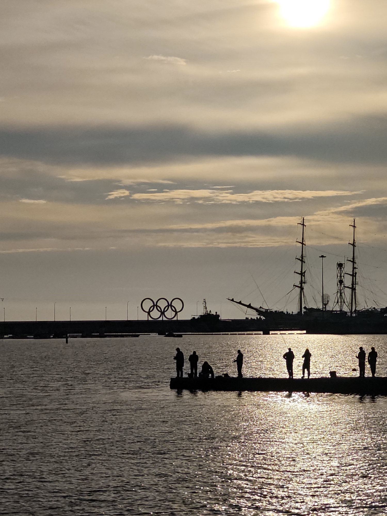 The image depicts a waterfront scene at sunset in Sochi, Russia. In the background are the Olympic rings, an embankment, and several ships. In the foreground, a group of people are standing and sitting along a pier, possibly fishing, as suggested by their postures. The sun is visible in the sky, casting a bright reflection on the water's surface. The overall lighting is muted, creating silhouettes of the people and objects.