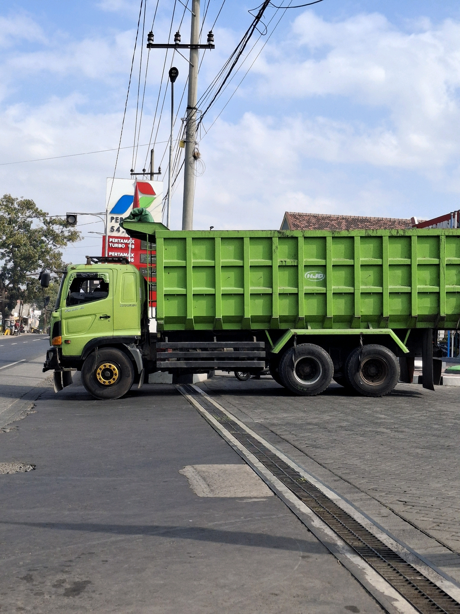 The image shows a bright green dump truck stopped on a paved road in Lumajang, Indonesia, at what appears to be a railway crossing. The truck is positioned on the right side of the frame, with its front cab on the left. The truck's bed is tall and segmented, with a "HJG" logo visible on its side. The front of the truck displays "HINO 500 FG 235 JD" badging. In the background, a Pertamina gas station sign is visible, displaying prices for "PERTAMAX TURBO" and "PERTAMAX." Utility poles with numerous wires crisscross the sky, which is partly cloudy. The road surface shows signs of wear, with cracks and patches. A metal grate runs along the edge of the road, adjacent to the truck. Buildings, trees, and other vehicles are visible in the distance along the road. The overall impression is an urban or semi-urban environment in daylight.