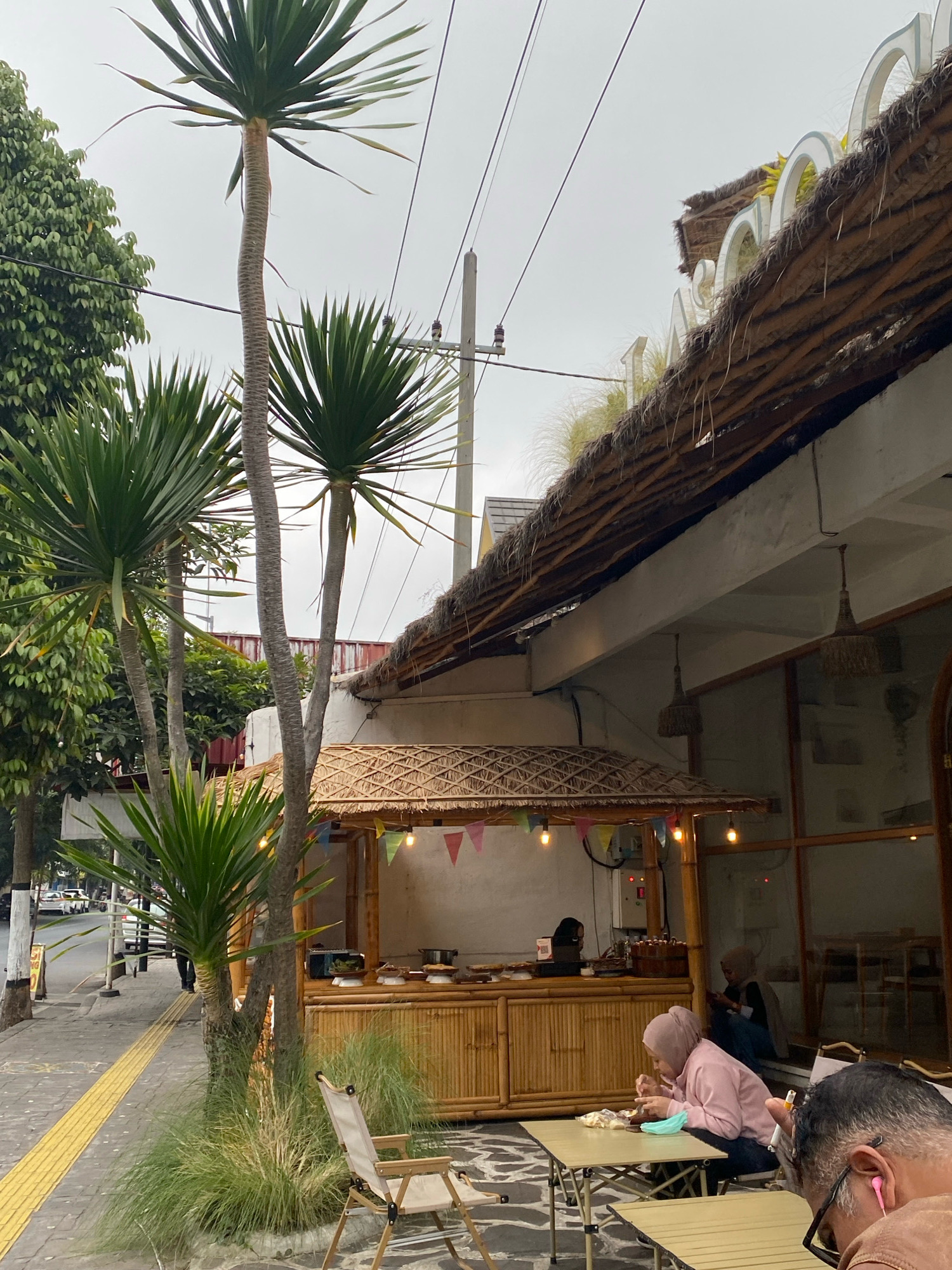 The image depicts an outdoor seating area of a cafe with a thatched roof and bamboo accents. In the foreground, a person in a pink hijab is seated at a light-colored table, eating. To their right, another person's head and shoulders are visible, also seated at a table. Behind them, in the cafe, another person in a hijab is seated. A bamboo food stall with colorful flags and lights is visible. Trees, including palm trees, line the sidewalk. The sidewalk has a yellow tactile paving strip. The sky is overcast. The setting appears to be a street in a city.