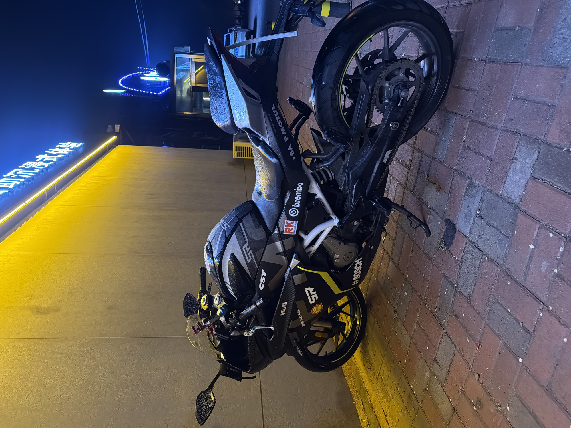 A black motorcycle is parked on a brick sidewalk in Zhongtun, China. The motorcycle has logos for CST, RK, Brembo, BA MOTUL, Shell Racing, and Bosch. The scene is illuminated by streetlights, casting a yellow glow on the adjacent building. A neon sign in Chinese characters is visible above the building. In the background, there is a blue neon structure, and the overall setting appears to be urban and nocturnal.