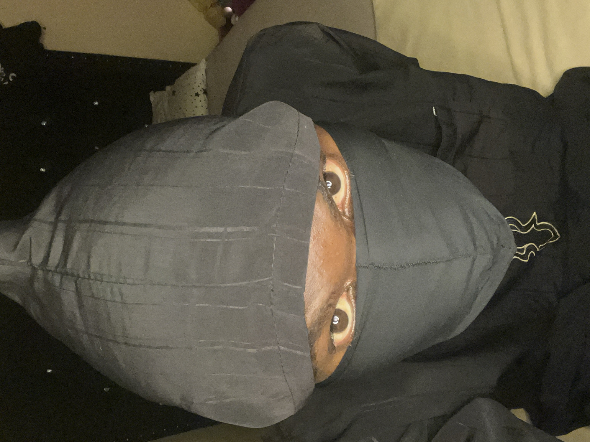 A close-up photograph captures a person lying down. Only their eyes and part of their nose and forehead are visible, peeking out from under a shiny grey durag. The person appears to be of African descent. They are lying on what appears to be a couch or bed, with black bedding to their right and an unseen headboard to the left. The photo is taken from an overhead angle, providing a clear view of the face.
The photograph does not provide any information about the person's location. Therefore, stating they are in Gura Top, Nigeria is not supported by the visual data.