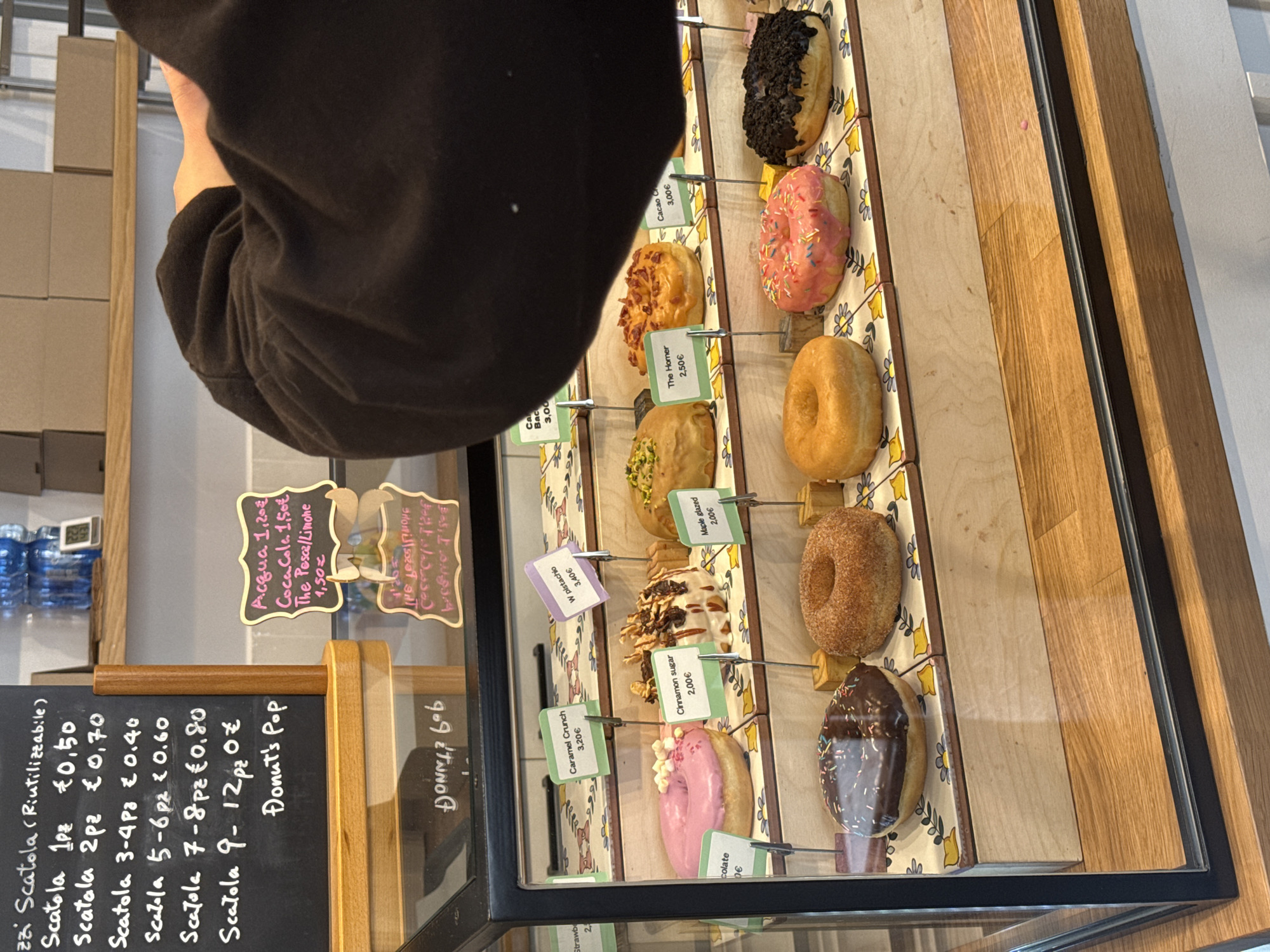 This image captures an overhead view inside a donut shop, likely in Milan, Italy, featuring a display case filled with a variety of donuts. The top left corner is partially obscured by a person's arm and sleeve, suggesting they might be interacting with the display or taking the photo.
The central focus is a multi-tiered display case showcasing numerous donuts, each presented on individual decorative trays with yellow and blue patterns. The donuts come in various flavors and toppings, including chocolate glazed with sprinkles, pink glazed with colorful sprinkles (one identified as "The Homer" for 2.50€), plain glazed, cinnamon sugar (2.00€), maple glazed (2.00€), pistachio (labeled "W pistachio" for 3.40€), and crunchy varieties like "Caramel Crunch" (3.20€) and "Cacao Crunch" (3.00€). A donut labeled "chocolate" is priced at 3.00€.
To the left of the display case, a black chalkboard lists prices for reusable boxes ("Scatola") based on the number of donuts: 1pc for 0.50€, 2pcs for 0.70€, 3-4pcs for 0.40€, 5-6pcs for 0.60€, 7-8pcs for 0.80€, and 9-12pcs for 1.00€. It also mentions "Donut's Pop". Another smaller sign on the right lists beverage prices: "Acqua 1,20€", "Coca Cola 1,50€", and "The Pesca/Limone 1,50€" (Peach/Lemon Tea). The prices are in Euros, and the Italian text confirms the location context. In the background, blue-capped water bottles are visible on a shelf. The setting appears clean and modern, with wooden accents in the display and surrounding furniture.