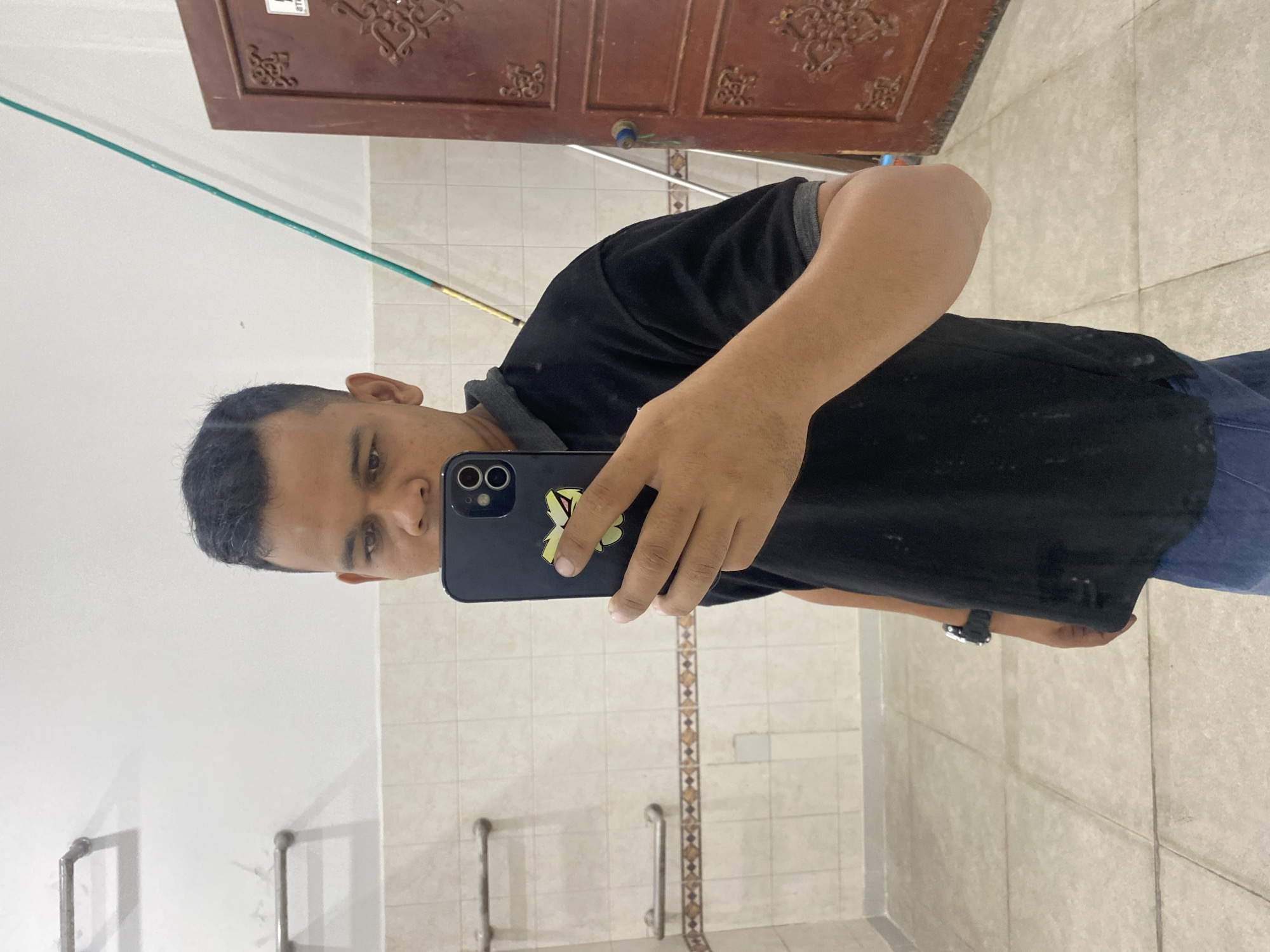 A man is taking a mirror selfie in a tiled room. He is wearing a black t-shirt and blue jeans. He is holding a smartphone in his right hand, which has a yellow sticker on the back. A green stick is leaning against the wall behind him. A dark red door with ornate carvings is visible to the right. The room appears to be a restroom or changing area with tiled walls and floor. There are two grab bars mounted on the tiled wall.