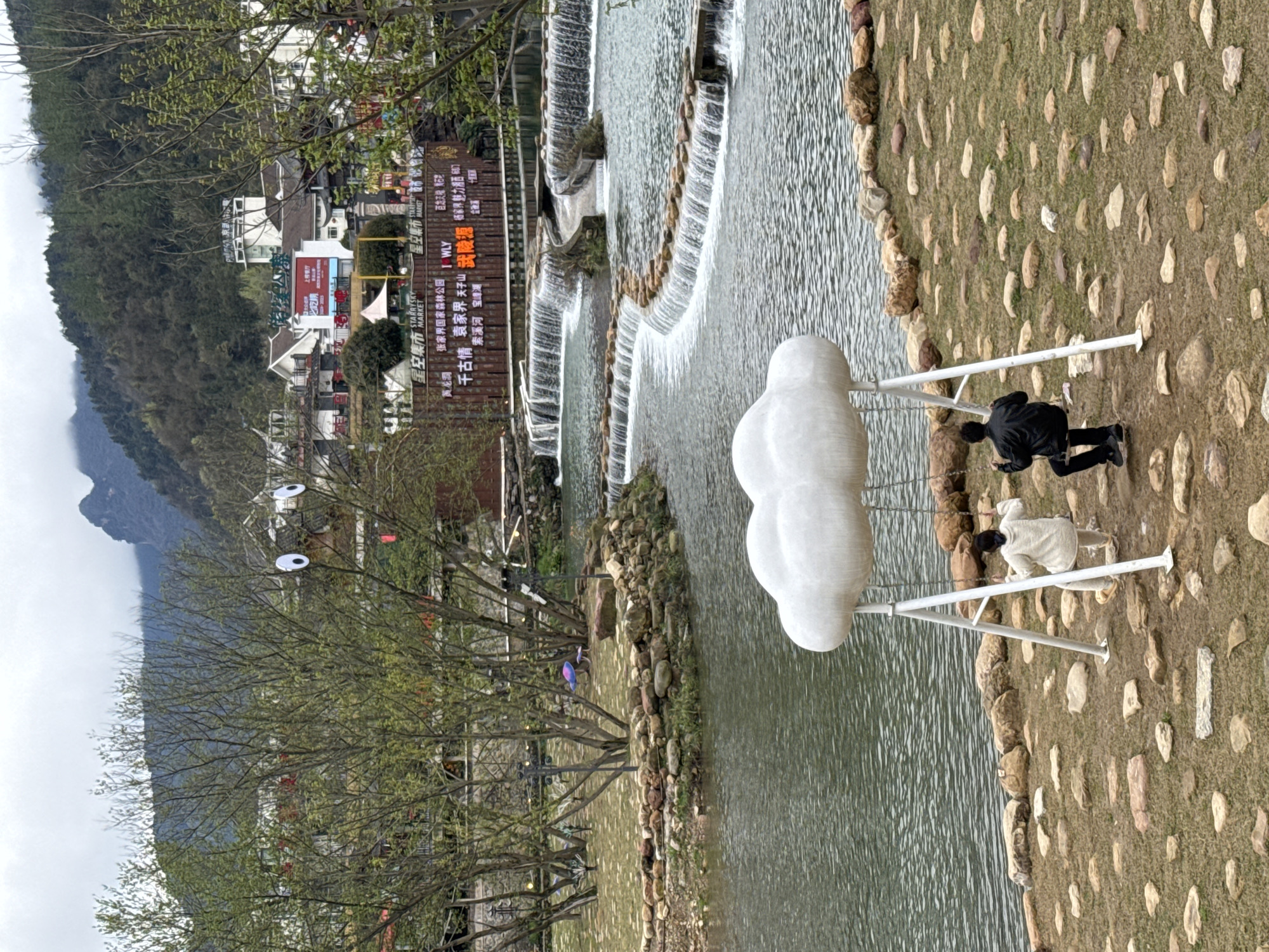 The scene depicts a landscaped recreational area within the city of Wulingyuan, China, characterized by a series of tiered waterfalls and surrounding parkland with architectural elements in the background.
In the foreground, two individuals are seated on a white, cloud-shaped swing structure positioned on a ground covered with sparse grass and embedded flat, irregular stones. The individual on the left swing seat is wearing light-colored, possibly off-white, casual clothing, including a long-sleeved top and pants. The individual on the right swing seat is dressed in a dark jacket and dark pants. Both individuals are facing away from the viewer, towards the water feature.
Immediately beyond the swing, a body of water, likely a stream or river, flows from the right towards the left. This water feature is characterized by a series of low, structured waterfalls or cascades, where water flows over several rock-lined, tiered levels. Rocky banks line the edges of the water.
Midground elements include several small, irregularly shaped islands or landscaped sections within or beside the water. These areas feature deciduous trees with budding leaves, some decorative lampposts, and colored mushroom-shaped sculptures. One prominent tree on a small island displays two large, white circular "googly eye" decorations attached to its upper branches.
In the background, a cluster of multi-story buildings, exhibiting a blend of traditional and contemporary Chinese architectural styles, rises behind the water features. These buildings include white facades and dark roofs. Several large commercial signs are visible; a prominent brown sign on the right displays "星空集市 STARRY SKY MARKET" in both Chinese and English, along with listings for local attractions such as "张家界国家森林公园" (Zhangjiajie National Forest Park), "天子山" (Tianzi Mountain), and "武陵源" (Wulingyuan). Another red sign indicates "上楼餐厅" (Upstairs Restaurant).
The furthest background shows densely forested mountains, some peaks partially obscured by clouds or mist, under an overcast sky. The overall environment appears to be a developed tourist destination.