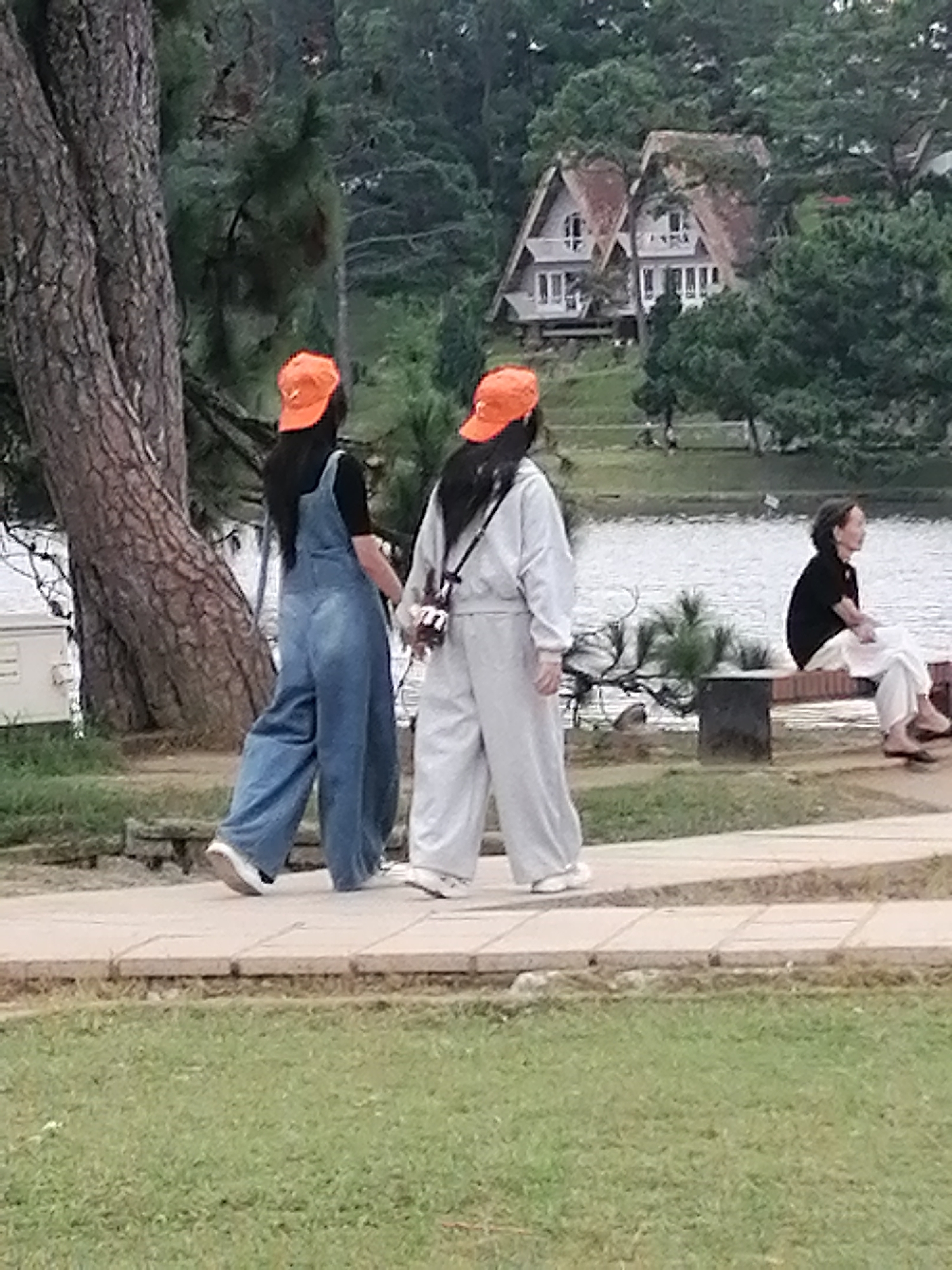 Two women, dressed in matching orange caps and loose-fitting outfits, walk side-by-side on a paved path near a body of water. One woman wears blue denim overalls, while the other wears a light grey tracksuit. Behind them, across the water, are two A-frame houses nestled among trees. To the right, a woman with short, dark hair sits on a bench, wearing a black top and white pants. The scene appears to be a park or scenic area, likely in Da Lat, Vietnam, known for its cool climate, pine forests, and French colonial architecture, which are reflected in the hilly landscape and the style of the houses.