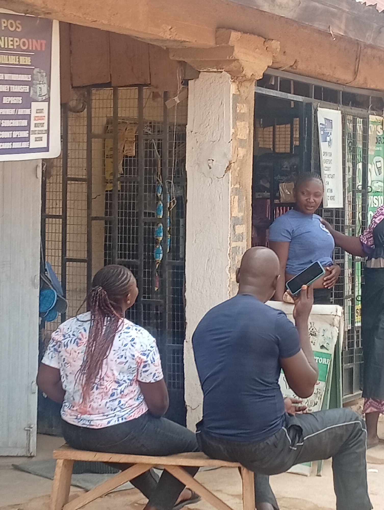 The image captures a street scene in Barakin Akawo, Nigeria. A woman with braids is sitting on a wooden bench, seen from the back, wearing a floral top and dark pants. A man is sitting next to her, also on the bench, with his back to the camera. He's holding a smartphone up in the air, possibly taking a picture. In the background, there's a shop front. A woman in a blue shirt stands in front of the shop. There are signboards visible near the entrance of the shop. The building appears to be made of concrete.