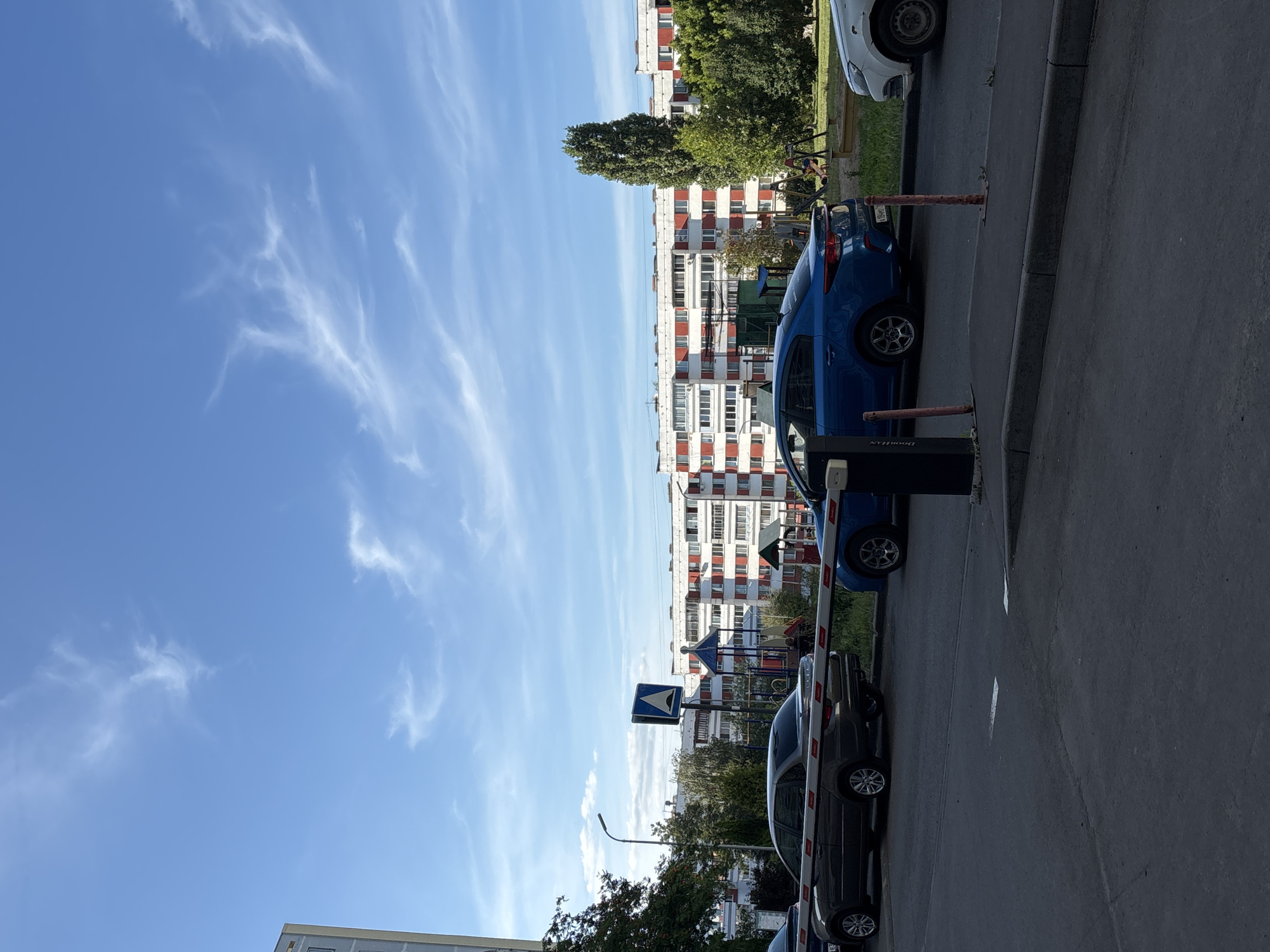 The image shows a street scene in Naberezhnye Chelny, Russia, on a clear day with scattered white clouds. In the foreground, a dark asphalt road is visible, with a gray sedan parked to the left and a blue sedan to the right, both partially obscured by a white barrier arm. Beyond the barrier, a playground with swings and a sandbox can be seen. In the background, a row of multi-story apartment buildings with red and white facades lines the street. A tall, slender tree stands to the right of the buildings. The overall impression is that of a residential area on a sunny day.