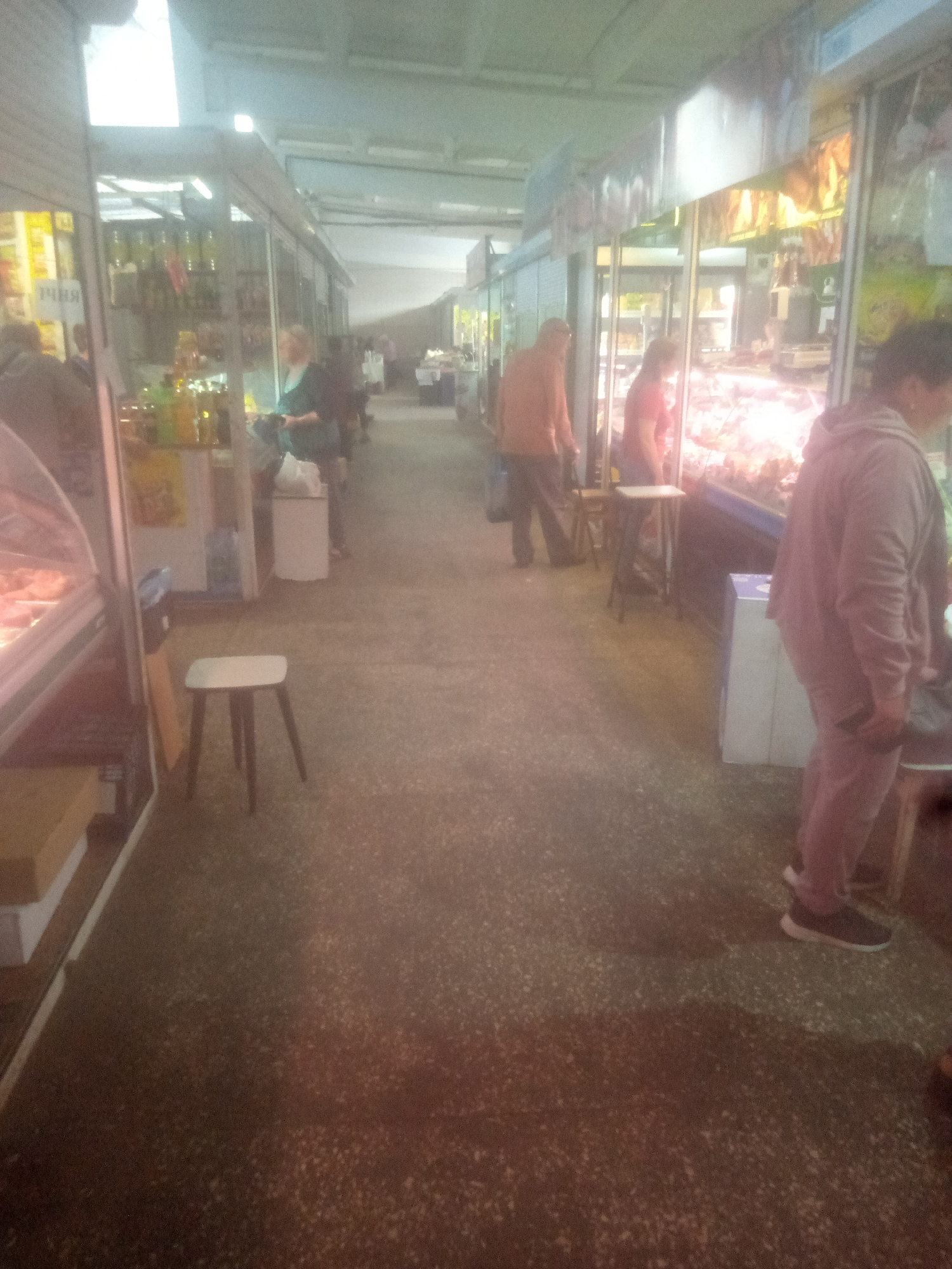 The image depicts a market scene with several stalls and people. In the foreground, a white stool with dark legs is positioned on a concrete floor. To the left, a glass display case containing food products is visible. Further down the market aisle, several individuals are present. One person, wearing a light-colored hooded jacket and dark pants, stands near a stall. Another person, in a red shirt, is behind a counter. A third person, in a pink hooded jacket and pants, is standing with their back to the camera, holding a phone. The stalls are lined up, with merchandise displayed in glass cabinets and on shelves. The overall environment appears to be an indoor market with artificial lighting.