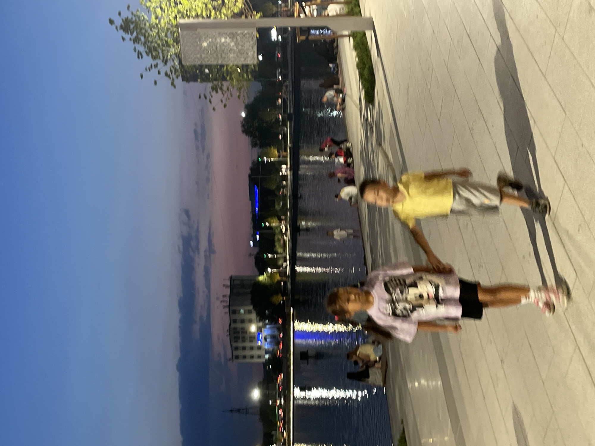 The image captures a twilight scene at the Almaty Central Park lake in Almaty, Kazakhstan. In the foreground, two children are walking along a paved promenade. A girl on the left, wearing a purple t-shirt with a cartoon character print and black shorts, is holding hands with a boy on the right, who is dressed in a yellow polo shirt and gray shorts. Both children are in motion, appearing slightly blurred due to movement. Behind them, the lake reflects the lights from the city and the sky, which displays a gradient of blue and pink hues, indicating sunset or twilight. Several other individuals are seated or standing along the edge of the lake in the middle ground, appearing as silhouettes against the water. In the background, buildings and streetlights are visible on the far side of the lake, with a tower in the distance. A modern structure with decorative paneling and a tree are positioned on the right side of the frame, closer to the foreground. The promenade is constructed with light-colored rectangular paving stones.