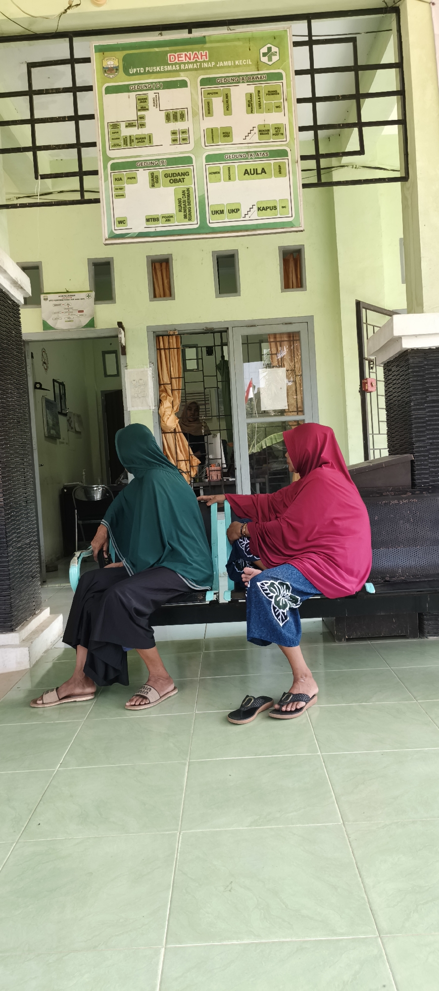 Two women are seated on a dark bench in front of a light green building with a tiled floor in Jambi Kecil, Indonesia. The woman on the left wears a dark green hijab and black pants. The woman on the right wears a maroon hijab and a blue patterned skirt. Both women are facing away from the camera, towards a doorway. Behind them, a sign displays a map of the UPTD Puskesmas Rawat Inap Jambi Kecil. The environment appears to be an outdoor waiting area or porch of a clinic.