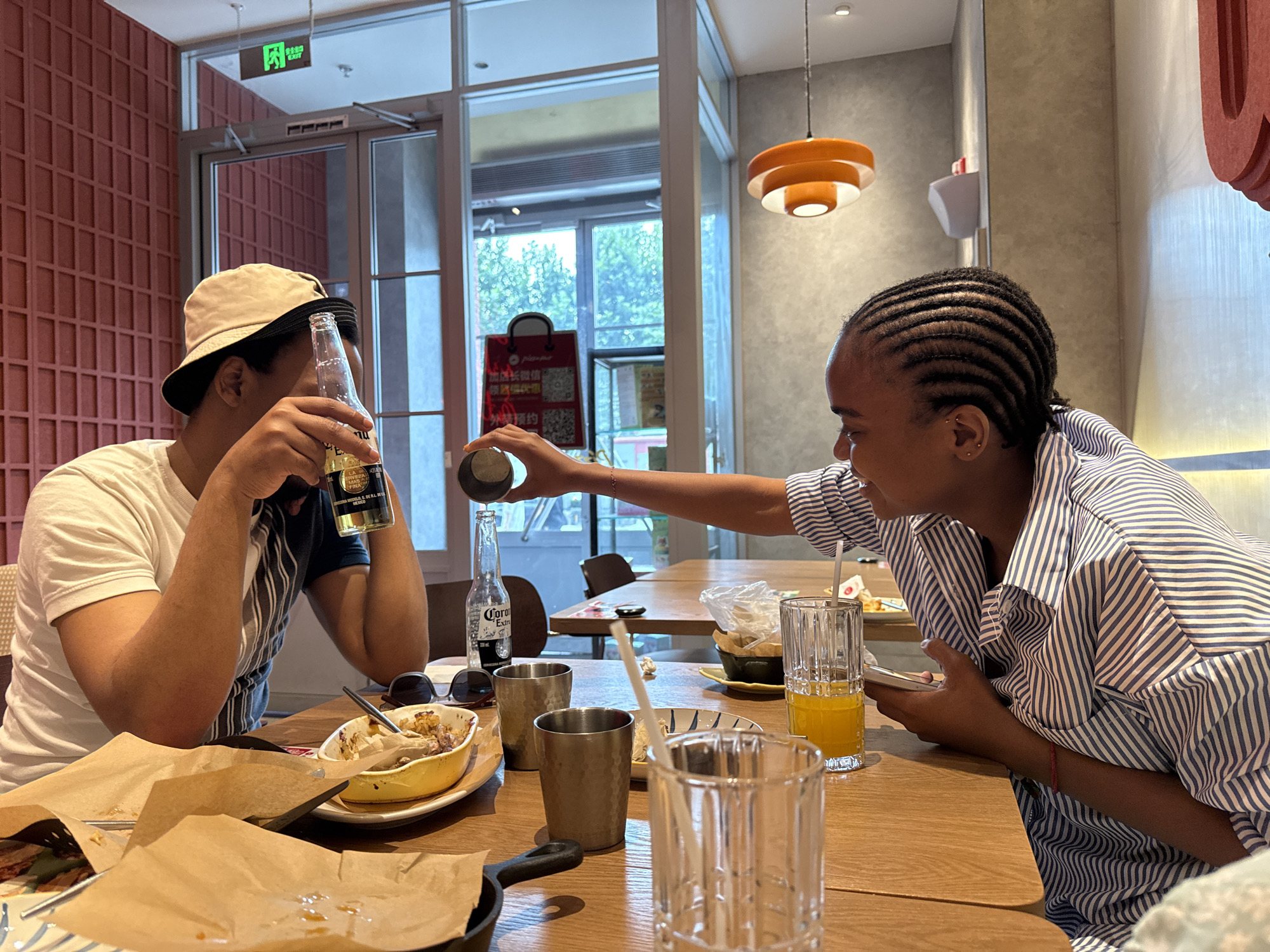 In Daxing District, China, two individuals are seated at a wooden table in a restaurant. The person on the left, wearing a white t-shirt and a beige bucket hat, holds a bottle of beer. The person on the right, with braided hair and wearing a blue and white striped shirt, is pouring liquid from a small metal cup into a bottle of beer. On the table are two glasses with orange liquid, several crumpled paper wrappers, a small yellow dish with food remnants, two metal cups, a pair of sunglasses, and a partially visible black cast-iron pan. The background features a large window with a view of trees, a red tiled wall, and modern light fixtures.